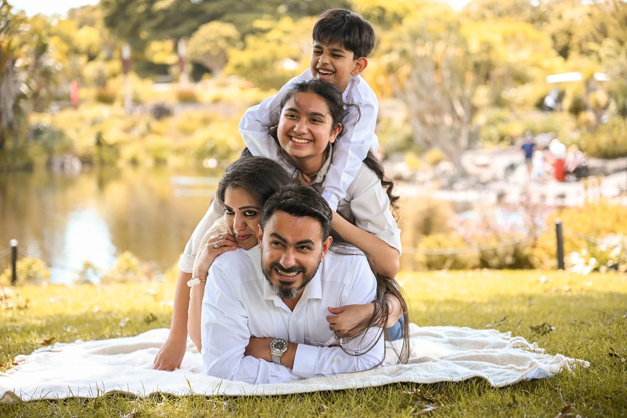 A family of four outdoors on a sunny day, stacked on top of each other on a blanket, smiling and enjoying nature.