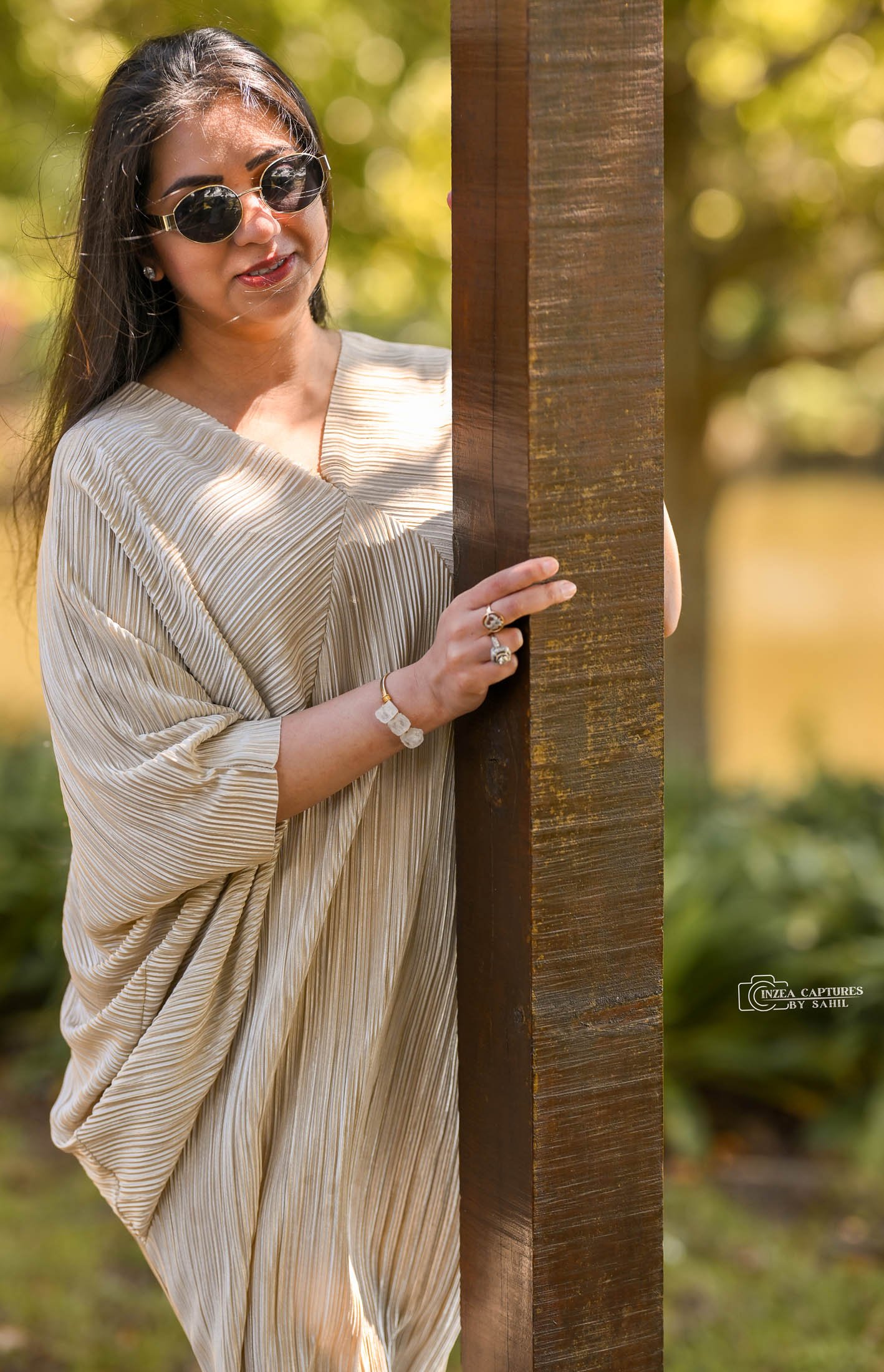 A woman wearing sunglasses and a beige pleated dress standing outdoors behind a wooden post, holding the edge of the post.
