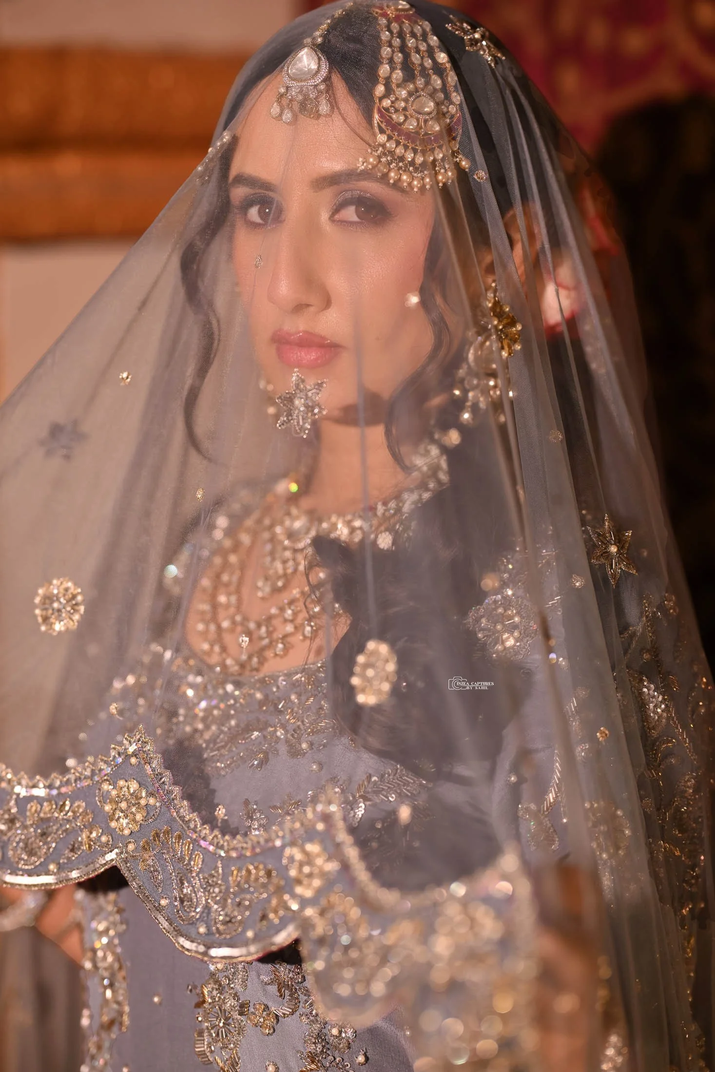 A woman wearing traditional bridal jewelry and clothing, with a sheer veil covering her face, adorned with intricate gold and silver embroidery and embellishments, and multiple earrings and necklaces.
