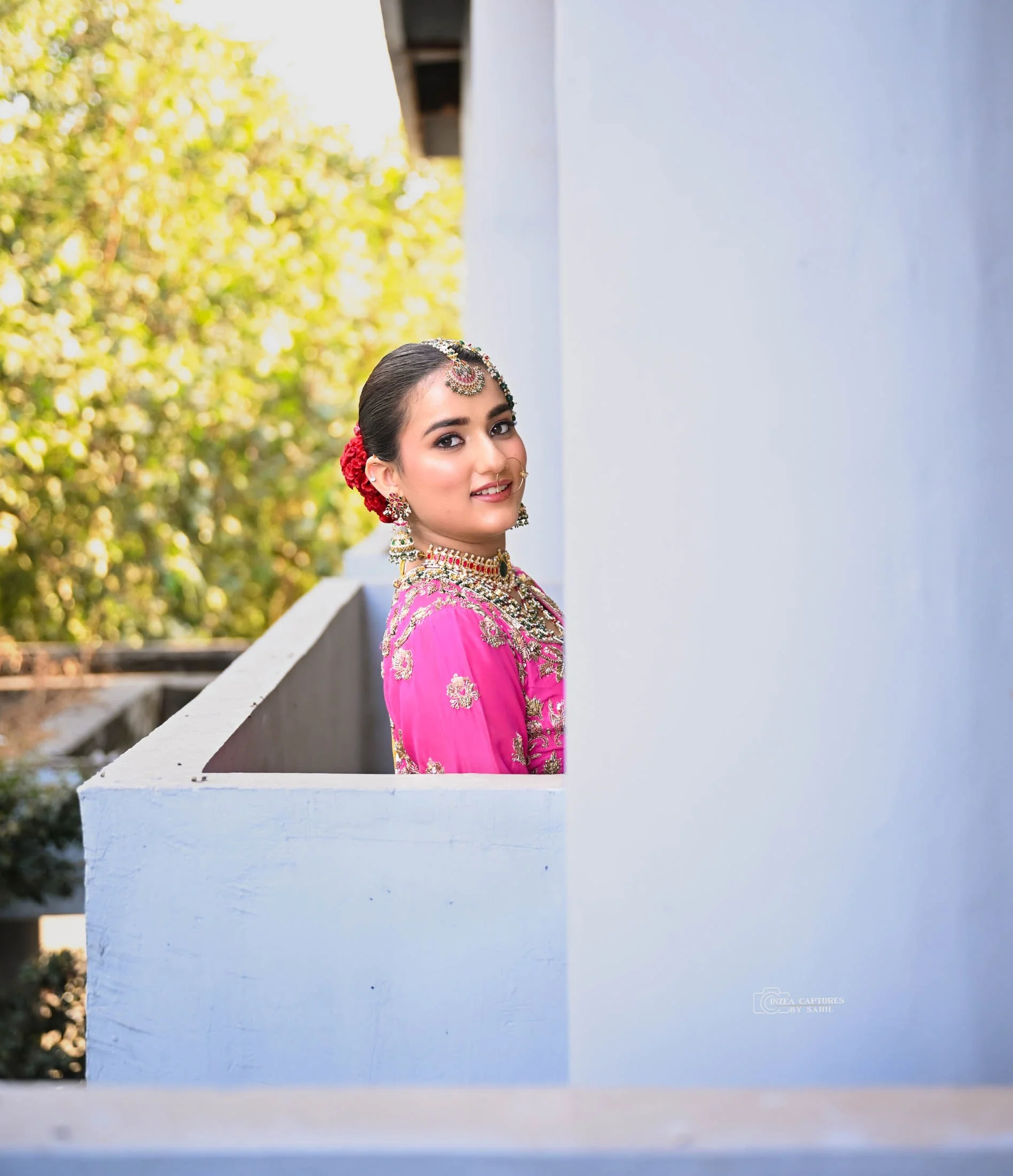 A woman wearing traditional Indian attire and jewelry, standing on a balcony with a white wall, with trees in the background.