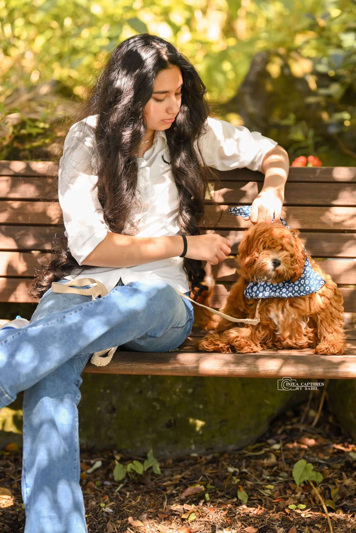 A young woman with long dark curly hair sitting on a park bench adjusting her adorable reddish-brown puppy's blue bandana. The scene is outdoors with lush green foliage and sunlight filtering through the trees.