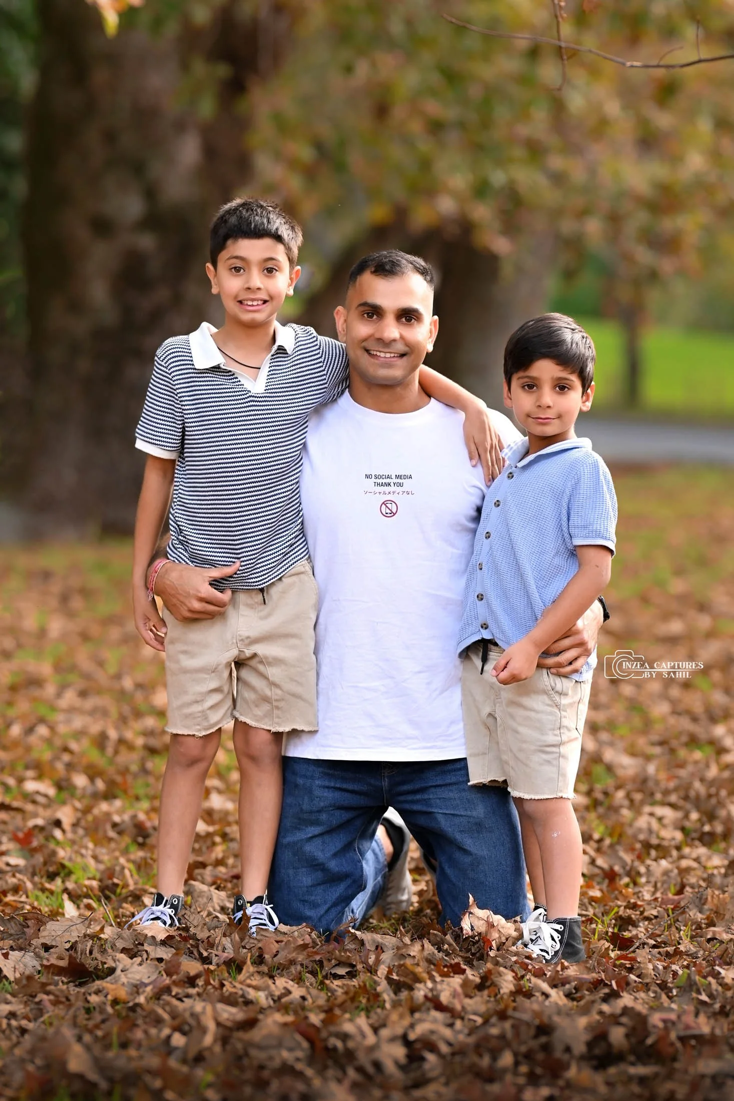 A man with two boys outdoors on a leaf-covered ground in fall, smiling at the camera, with trees and a path in the background.