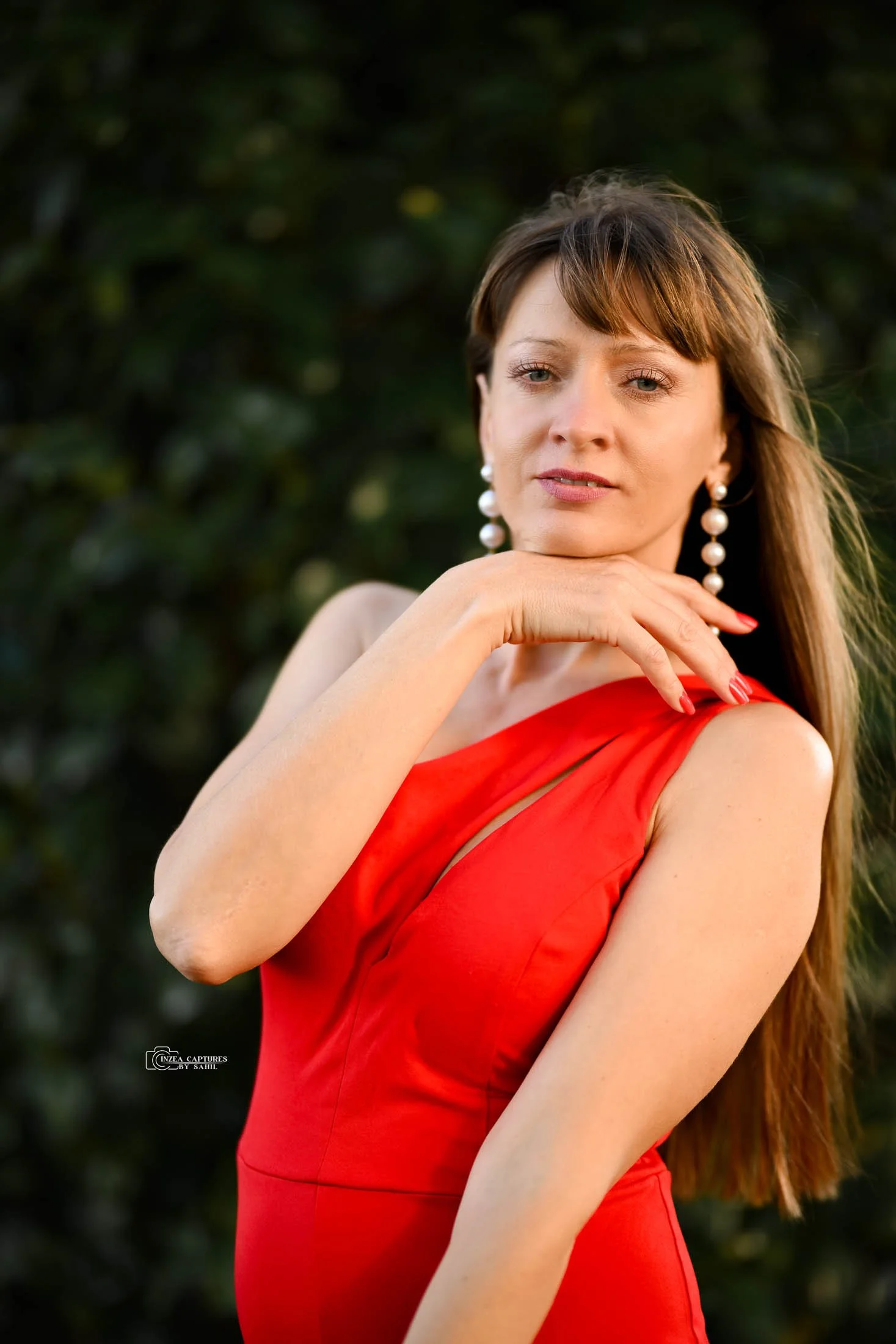 A woman in a red dress with pearl earrings, outdoors, with dark green foliage in the background, posing with her hand on her shoulder.