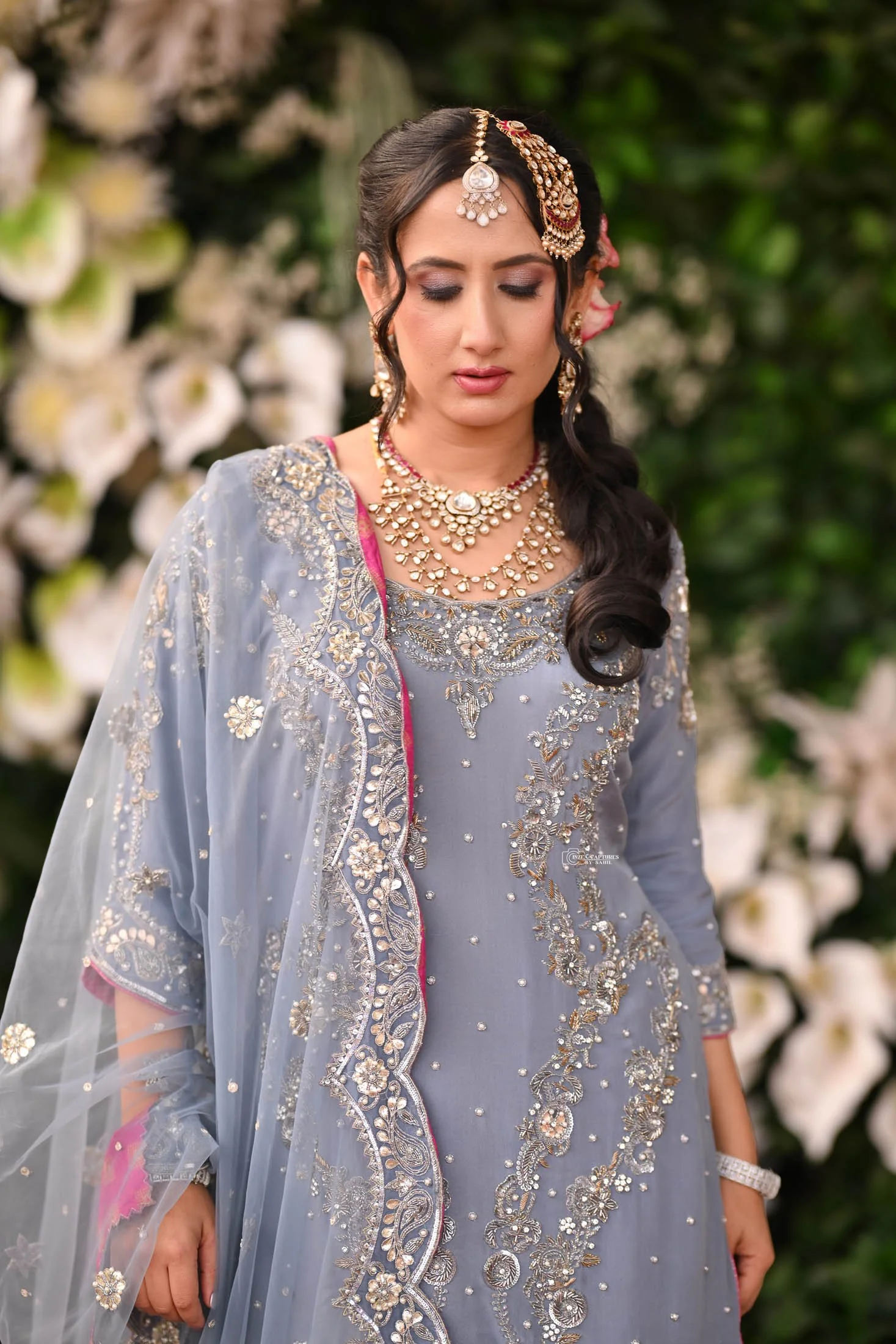 Woman dressed in traditional ornate South Asian attire with gold jewelry, standing against a backdrop of white flowers and green foliage.