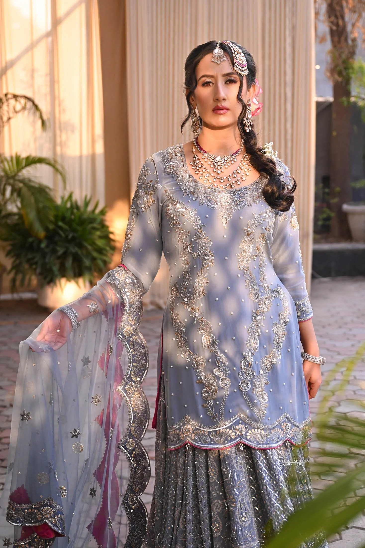Woman wearing traditional South Asian attire with intricate embroidery, jewelry, and a dupatta, standing outdoors.