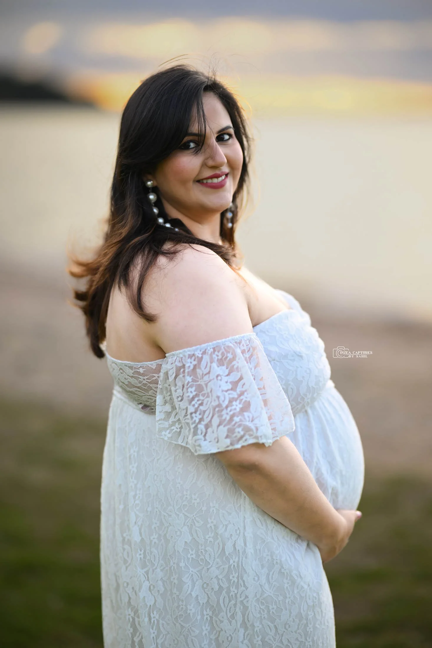 Pregnant woman outdoors at sunset, wearing a white lace off-shoulder dress and pearl earrings, smiling at camera.