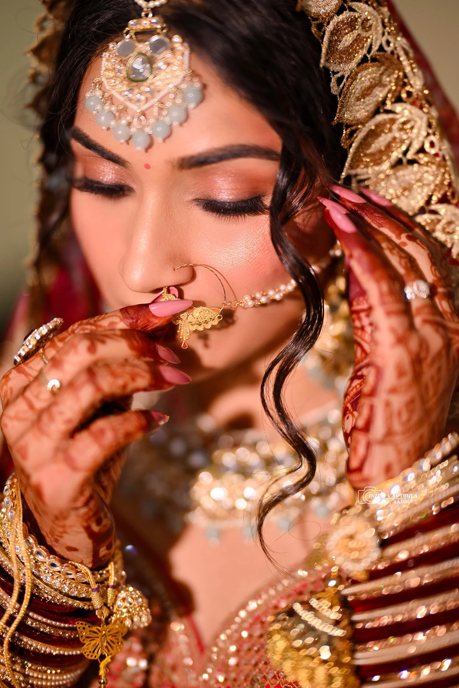 Close-up of an Indian bride wearing traditional jewelry and henna on her hands, with her eyes closed and holding a nose ring to her face.