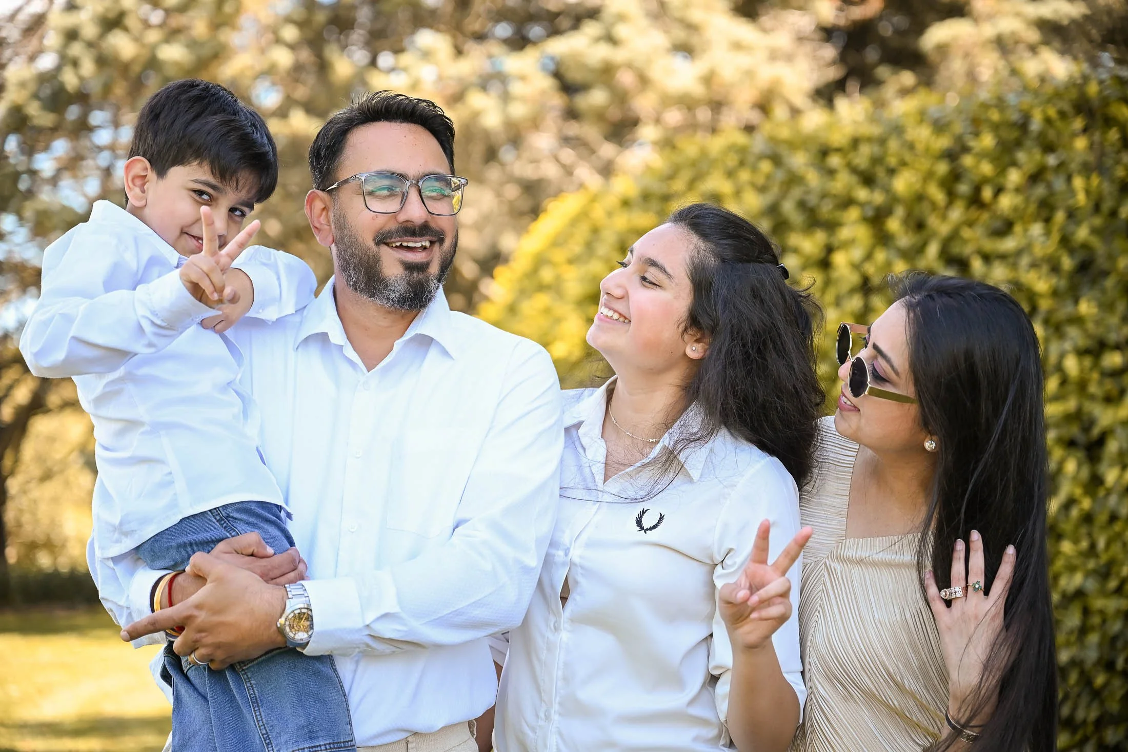 A group of four people smiling and enjoying outdoors in a park with trees in the background. A man is holding a young boy, and two women are standing nearby. One woman is making a peace sign with her fingers.