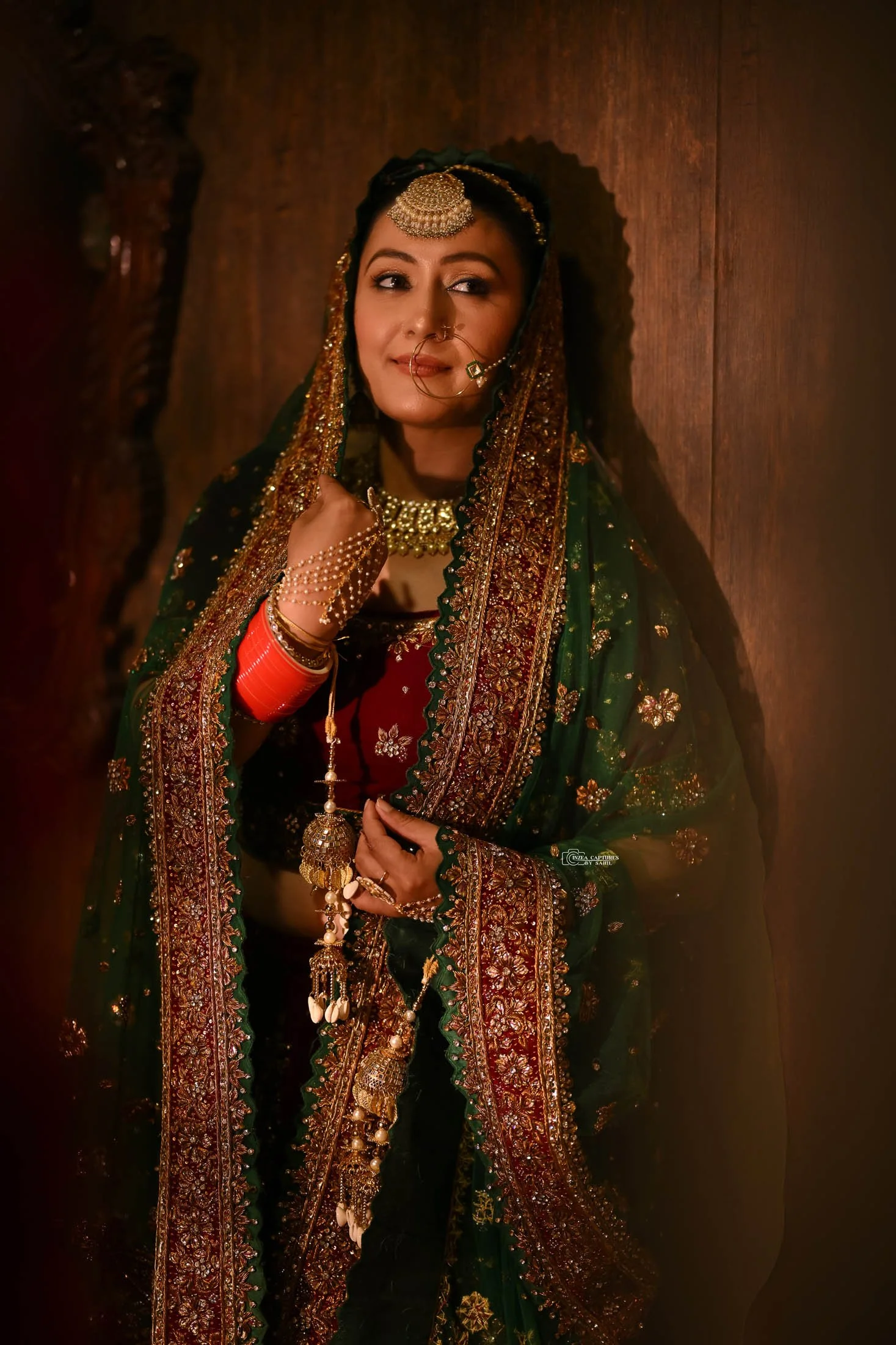 A woman dressed in traditional Indian attire, wearing ornate jewelry including a headpiece, nose ring, necklace, bangles, and a heavily embroidered shawl, stands against a wooden background.