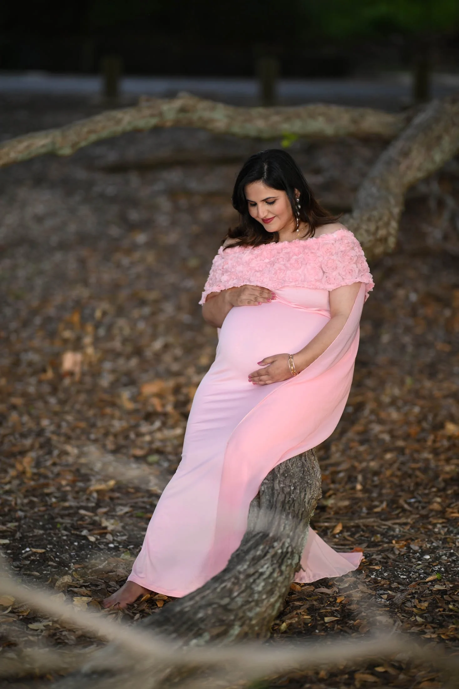A pregnant woman in a pink dress with a floral off-the-shoulder top, sitting on a tree branch in a wooded area, looking down at her belly.