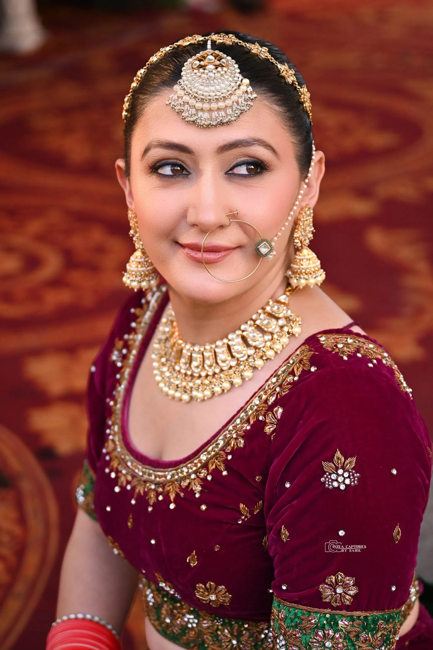 A woman dressed in traditional Indian attire with elaborate gold jewelry, including a headpiece, earrings, necklace, nose ring, and bangles.