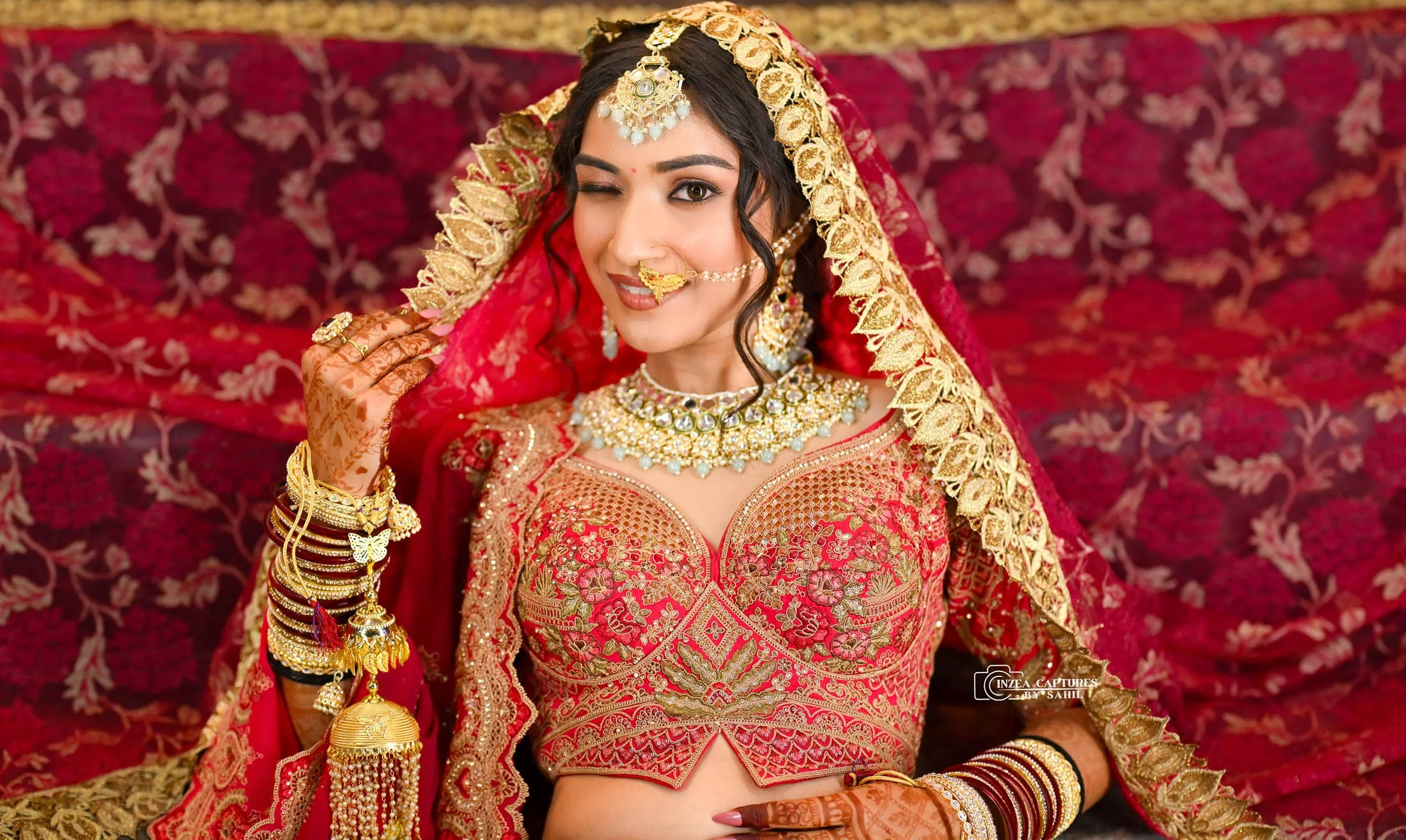 A woman in traditional Indian bridal attire, adorned with gold jewelry, including a necklace, earrings, nose ring, bangles, and headpiece, wearing a red and gold embroidered lehenga and veil, posing against a matching red and gold backdrop.
