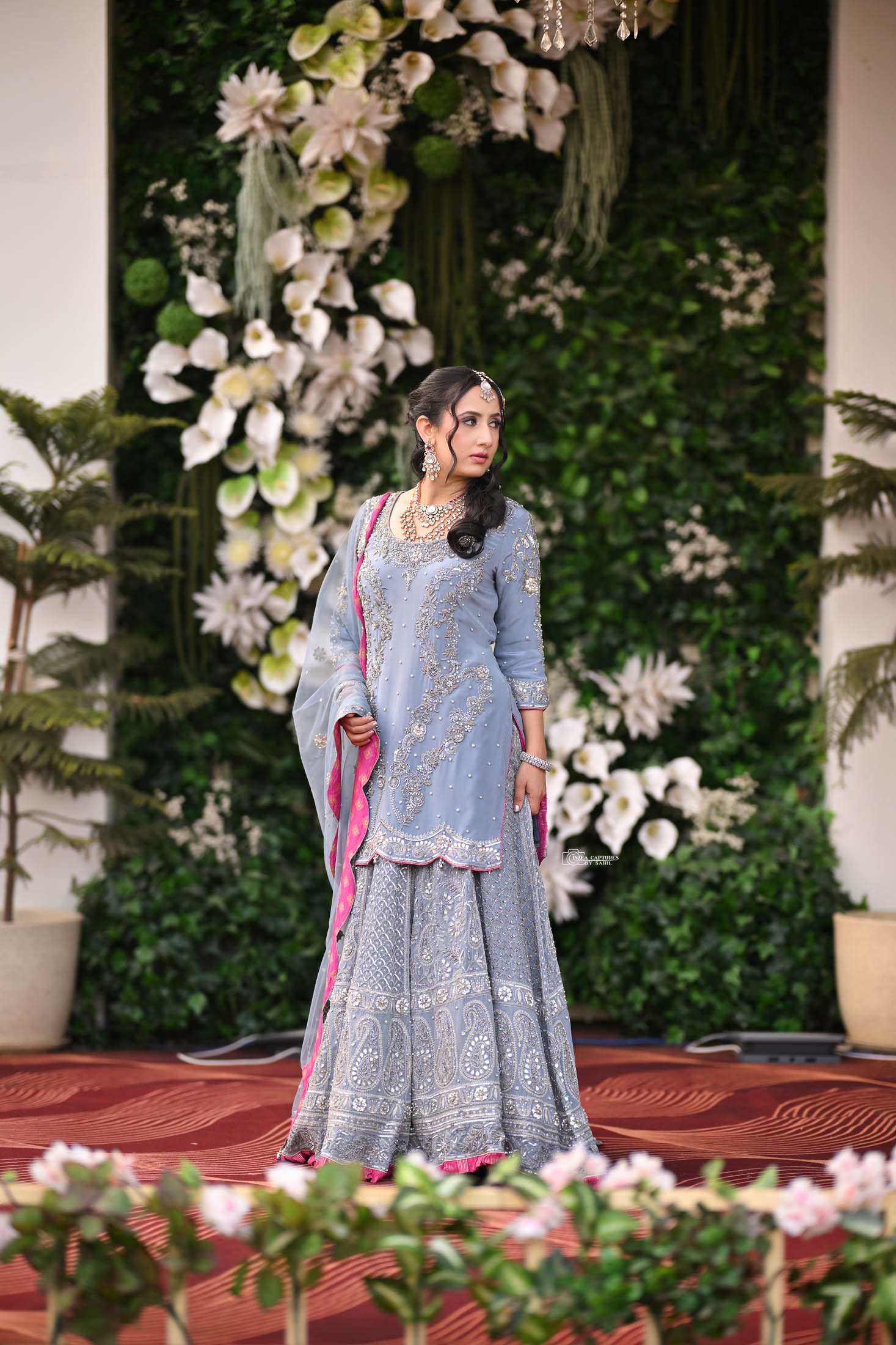 A woman in a traditional light blue embroidered dress with pink and silver accents standing on a decorated stage with a floral backdrop and potted plants.