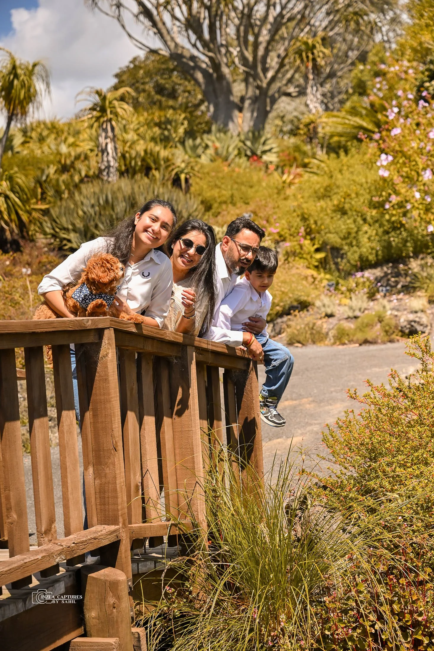 A family of five, including a woman, man, two children, and a dog, leaning on a wooden railing outdoors in a garden or park with lush greenery and colorful flowering plants, under a partly cloudy sky.