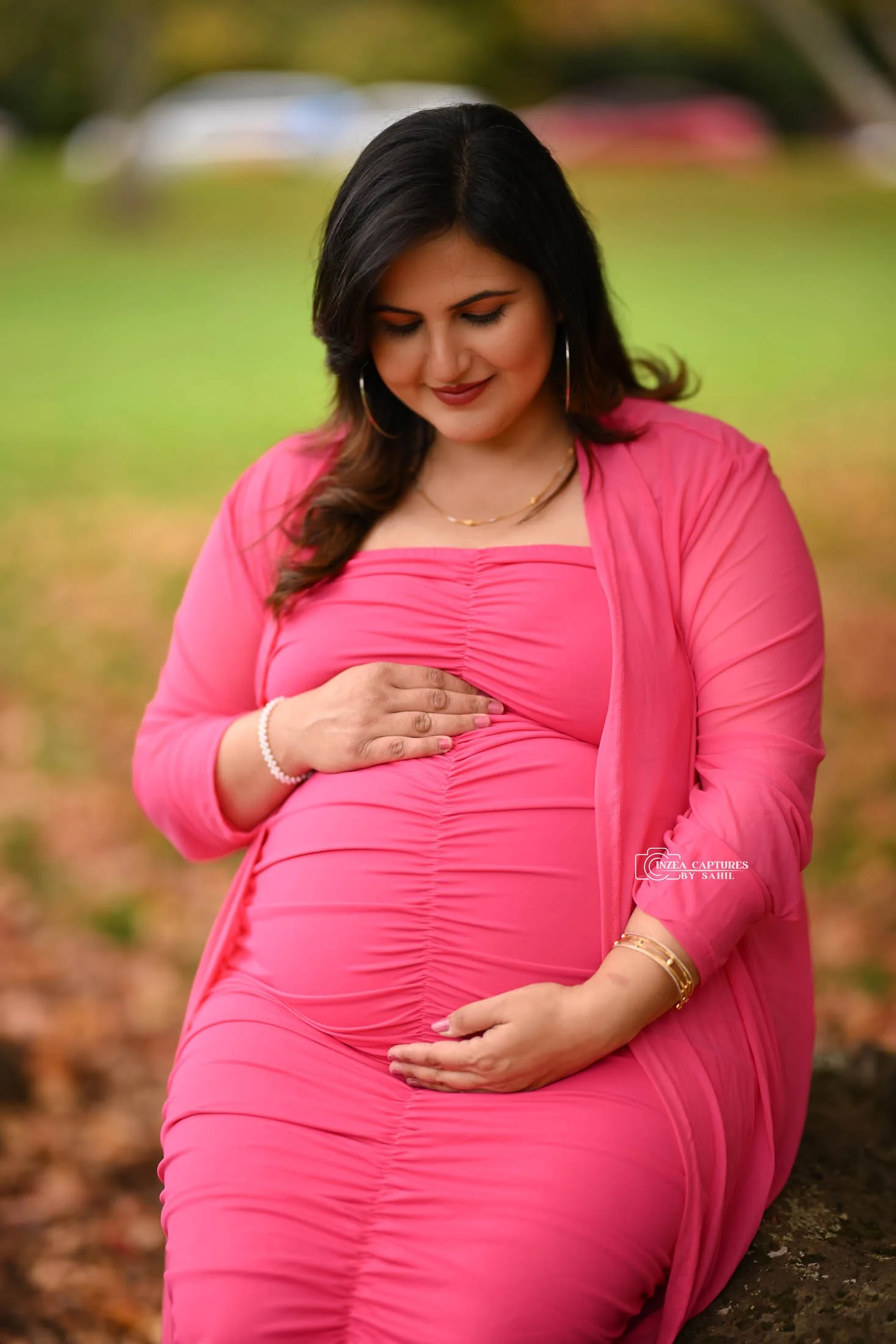 Pregnant woman in pink dress sitting outdoors, cradling her belly, with a blurred autumn background.