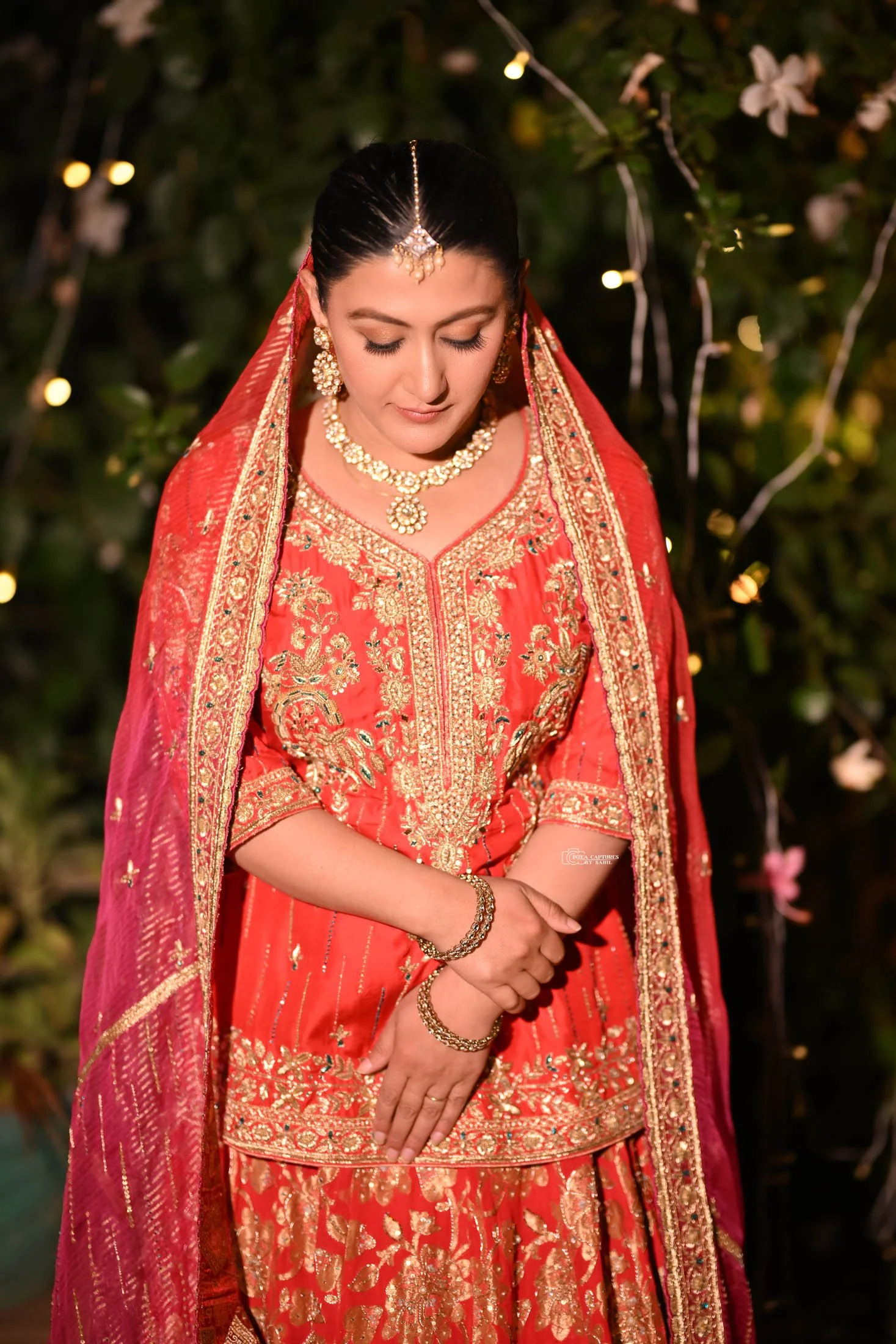 A woman dressed in a red traditional Indian outfit with gold embroidery, wearing jewelry and a veil, standing outdoors with string lights and flowers in the background.