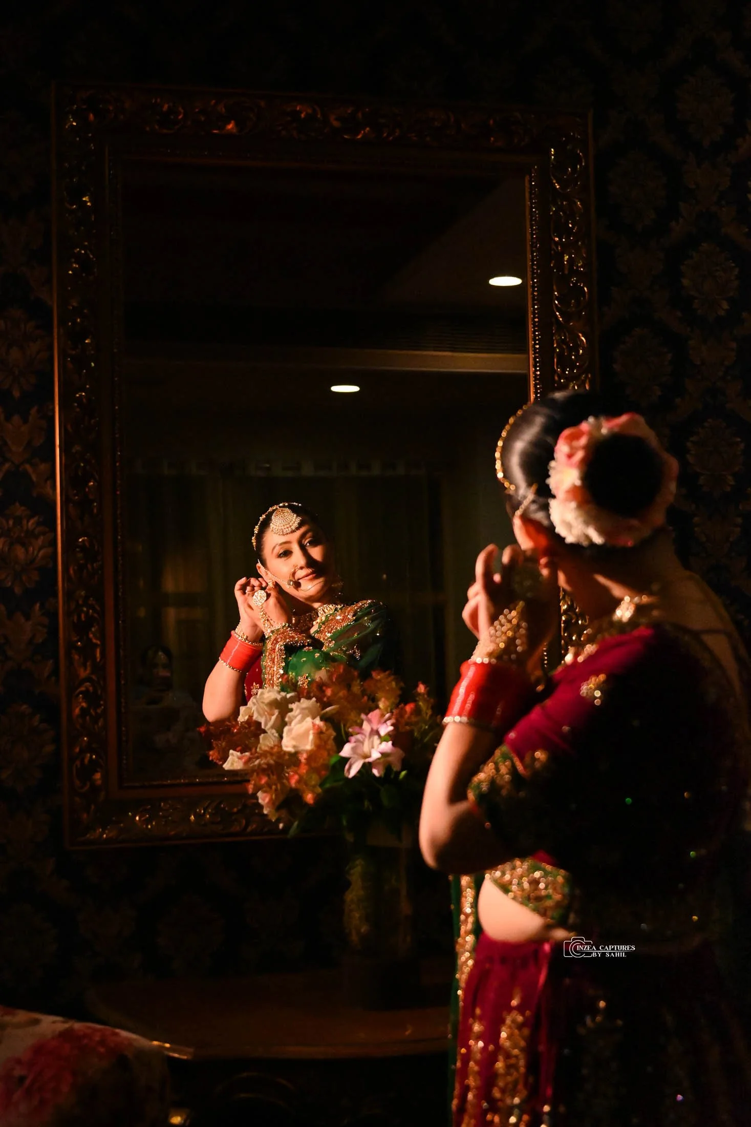 Indian woman in traditional attire looking at her reflection in a mirror, adjusting jewelry, with flowers on the table beside her.