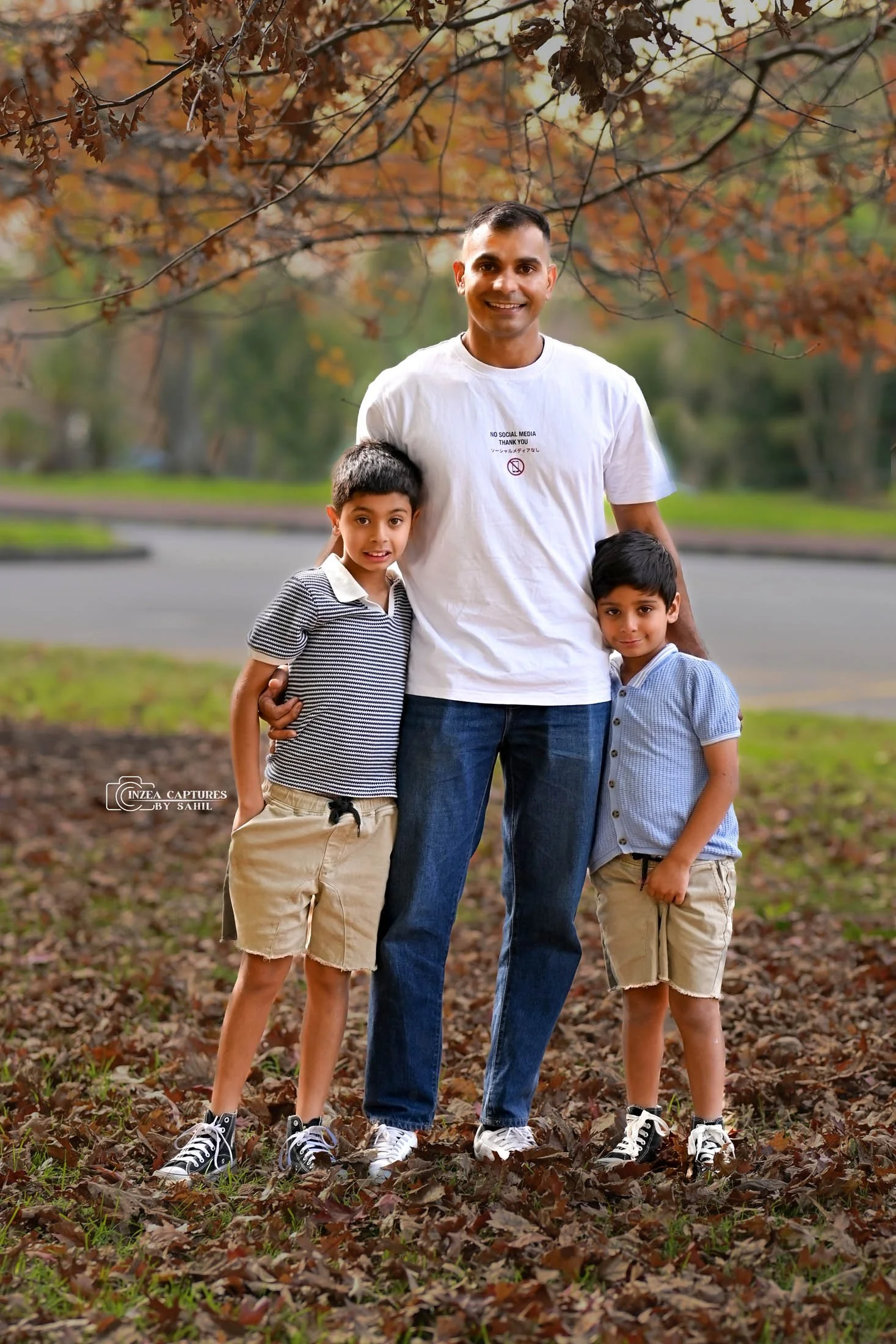 A man standing outdoors with two young boys, under a tree with autumn leaves, smiling at the camera.