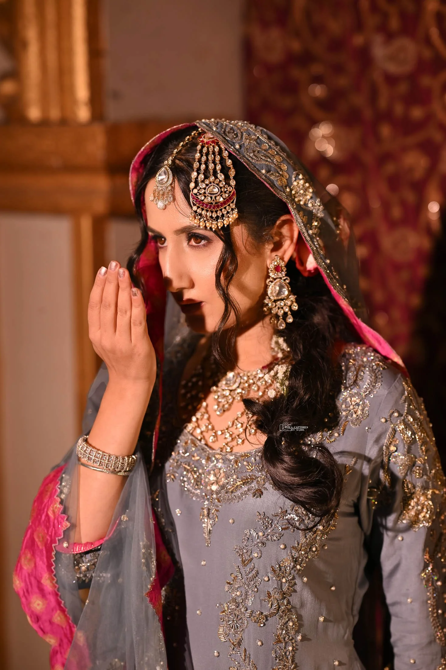 A woman dressed in traditional Indian attire and jewelry, with a detailed embroidery dress, wearing a headscarf, earrings, necklace, and a bracelet, touching her face with her hand.