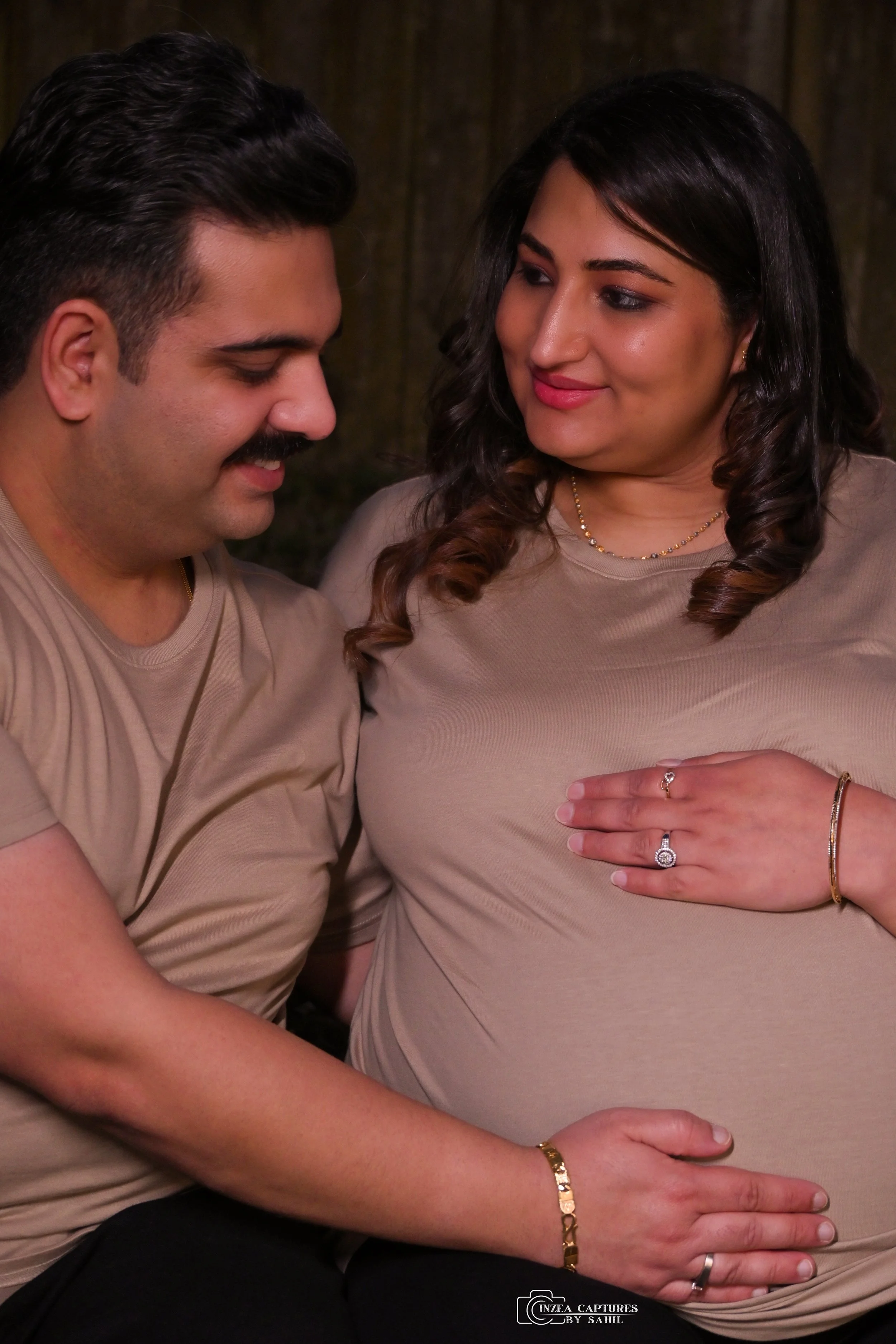 A pregnant woman and her partner are looking at her baby bump, both smiling gently, in a warm, intimate moment.