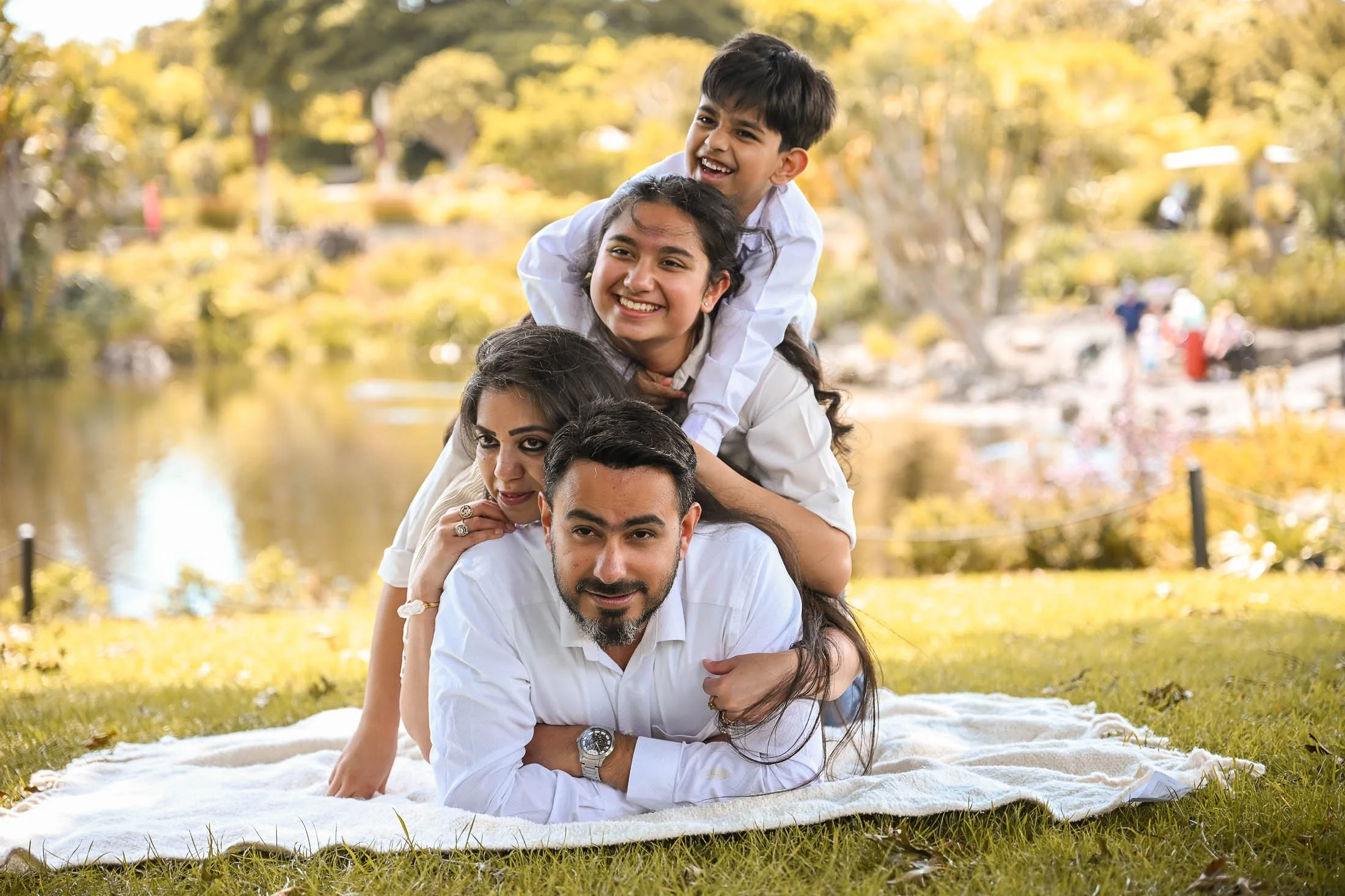 A family of four lying on a blanket in a park during fall, with trees and a pond in the background, enjoying the outdoor setting.