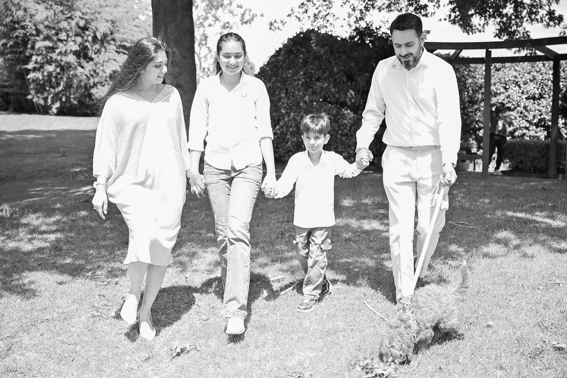 A family walking with a dog in a park, holding hands, with trees and a gazebo in the background.