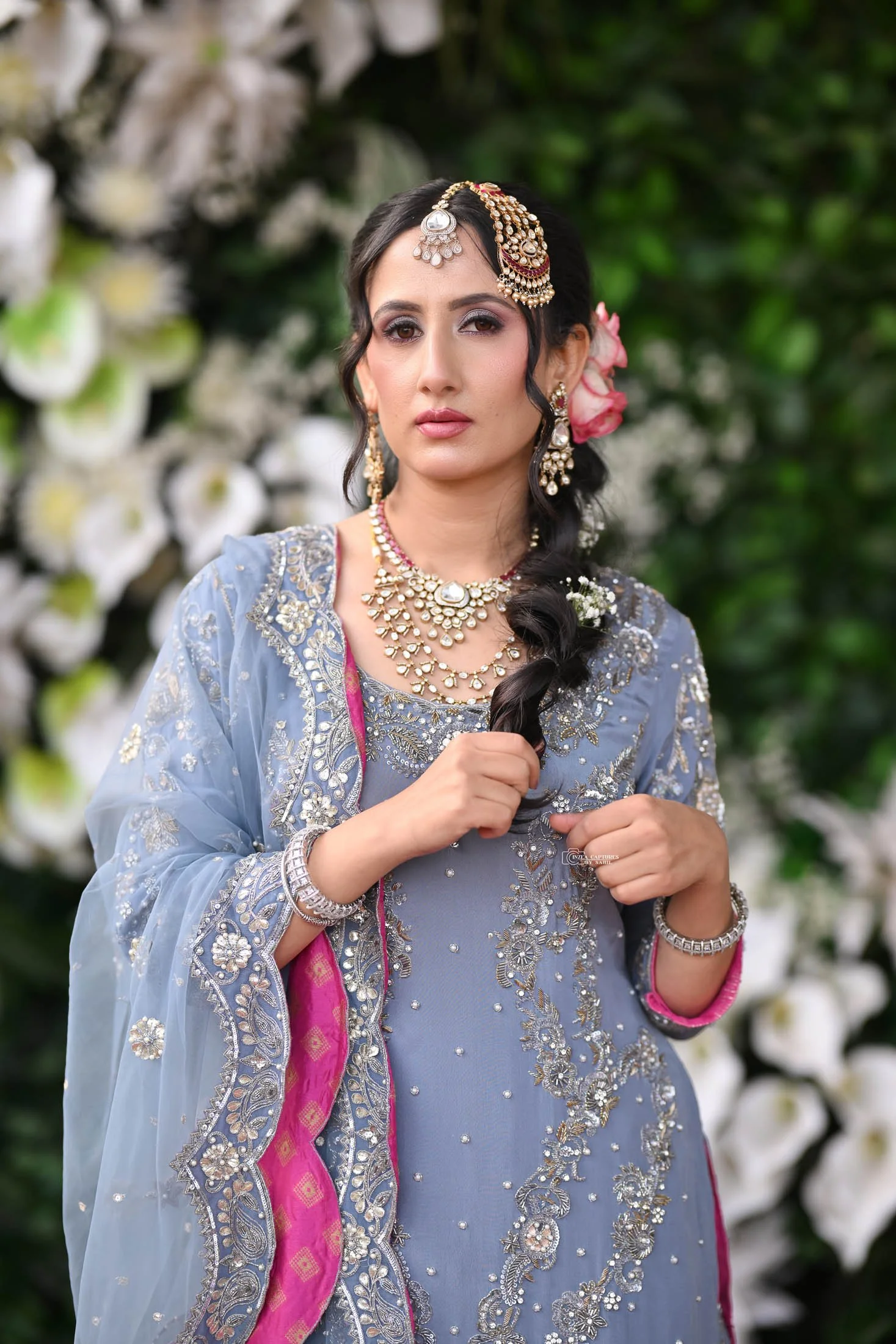 A woman wearing traditional Indian attire, jewelry, and a braid hairstyle, standing outdoors with a floral background.