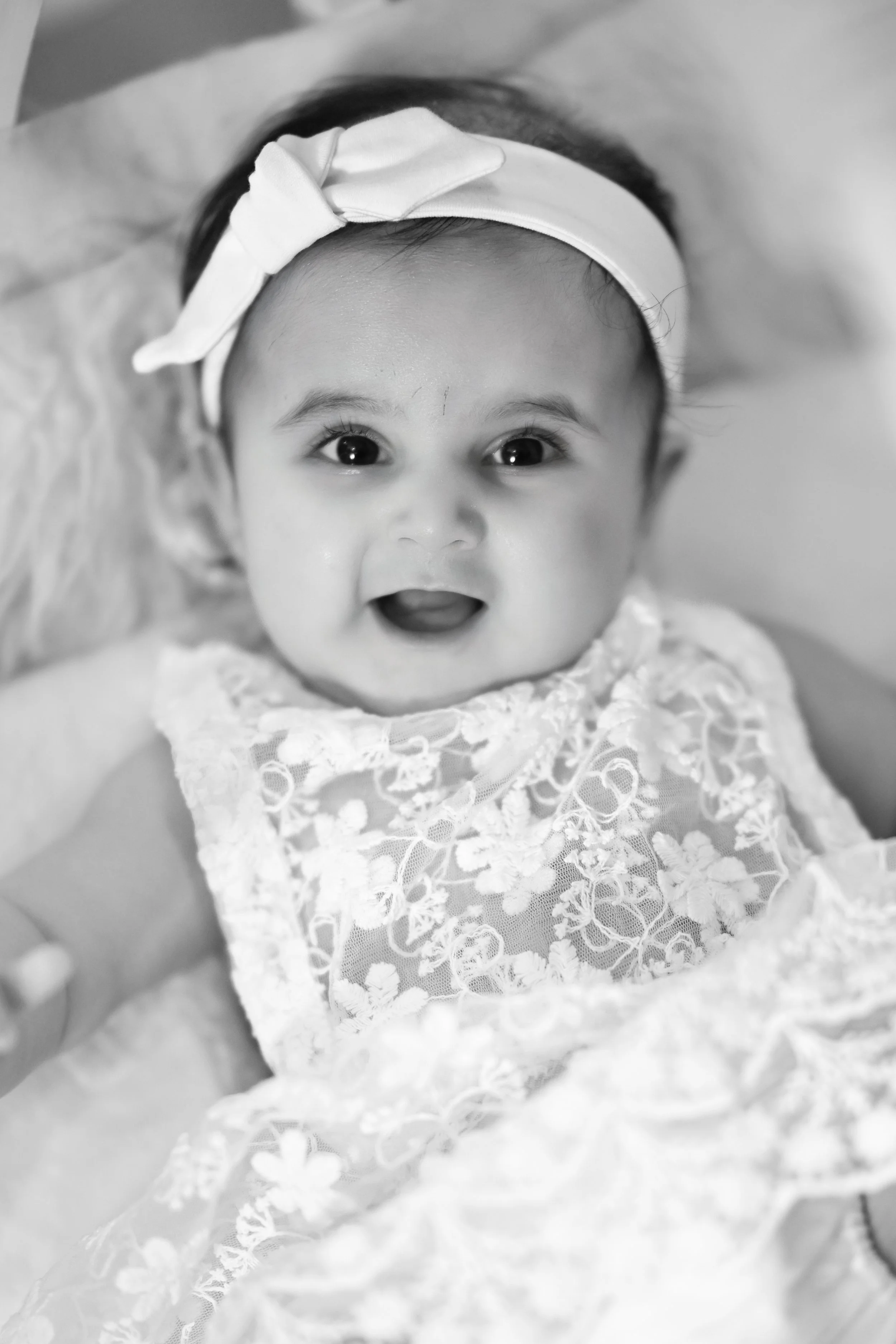 A baby girl with a bow headband, smiling and wearing a lace dress, lying on her back.