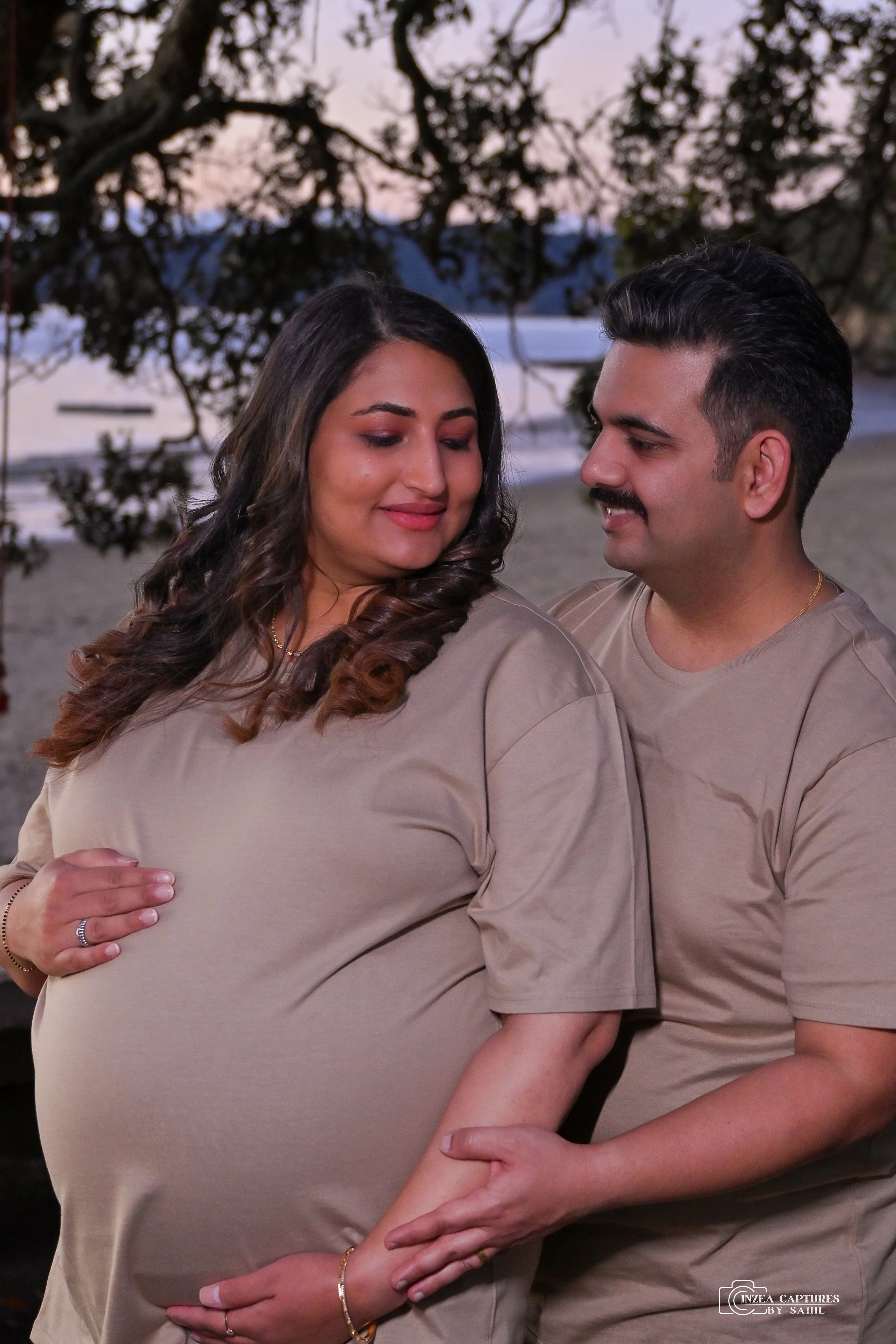 A pregnant woman and a man stand close together outdoors by the water at sunset, both wearing beige shirts. The woman has dark, curled hair and is smiling gently with her eyes closed, while the man looks at her fondly. The woman's hands are on her be