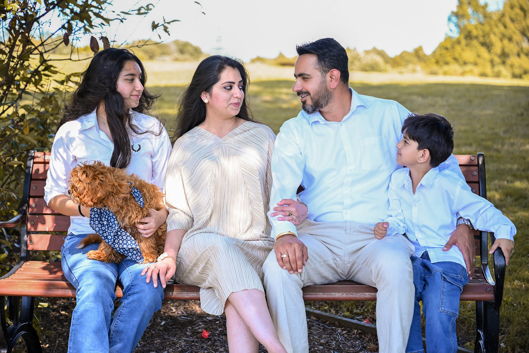 A family of five sitting on a park bench outdoors with trees and grass in the background, including a woman, man, two children, and a small dog.