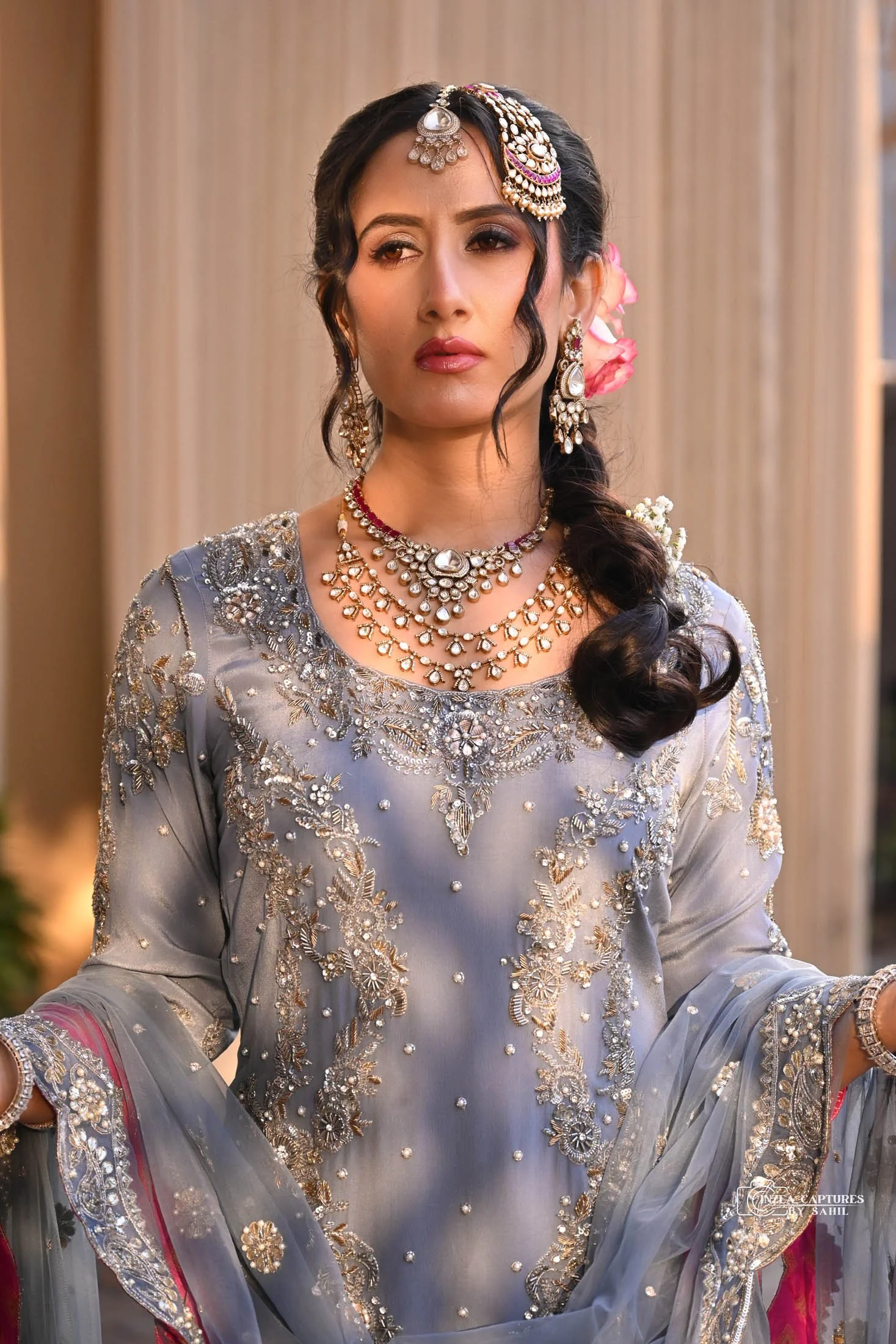 A woman dressed in an ornate silver and gold embroidered traditional Indian outfit, adorned with heavy jewelry including necklaces, earrings, and forehead jewelry, with a serious expression.