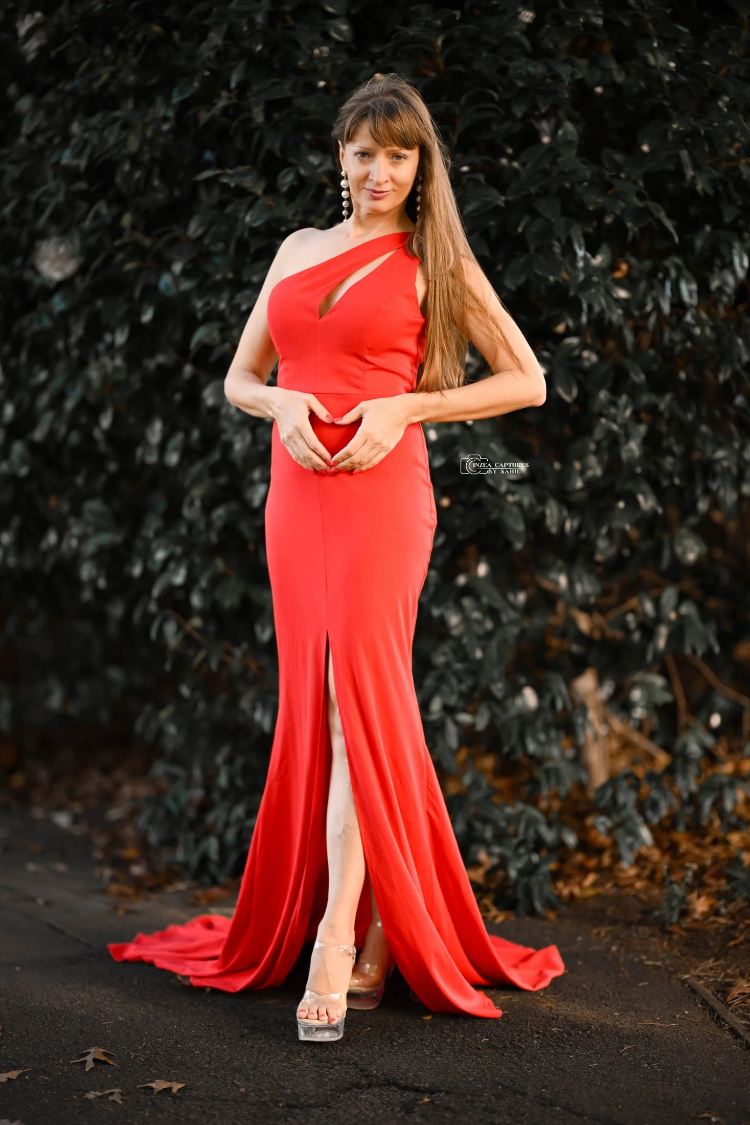 Woman in a red gown with a slit, standing outdoors on a paved path, with dark foliage in the background, making a heart shape with her hands.