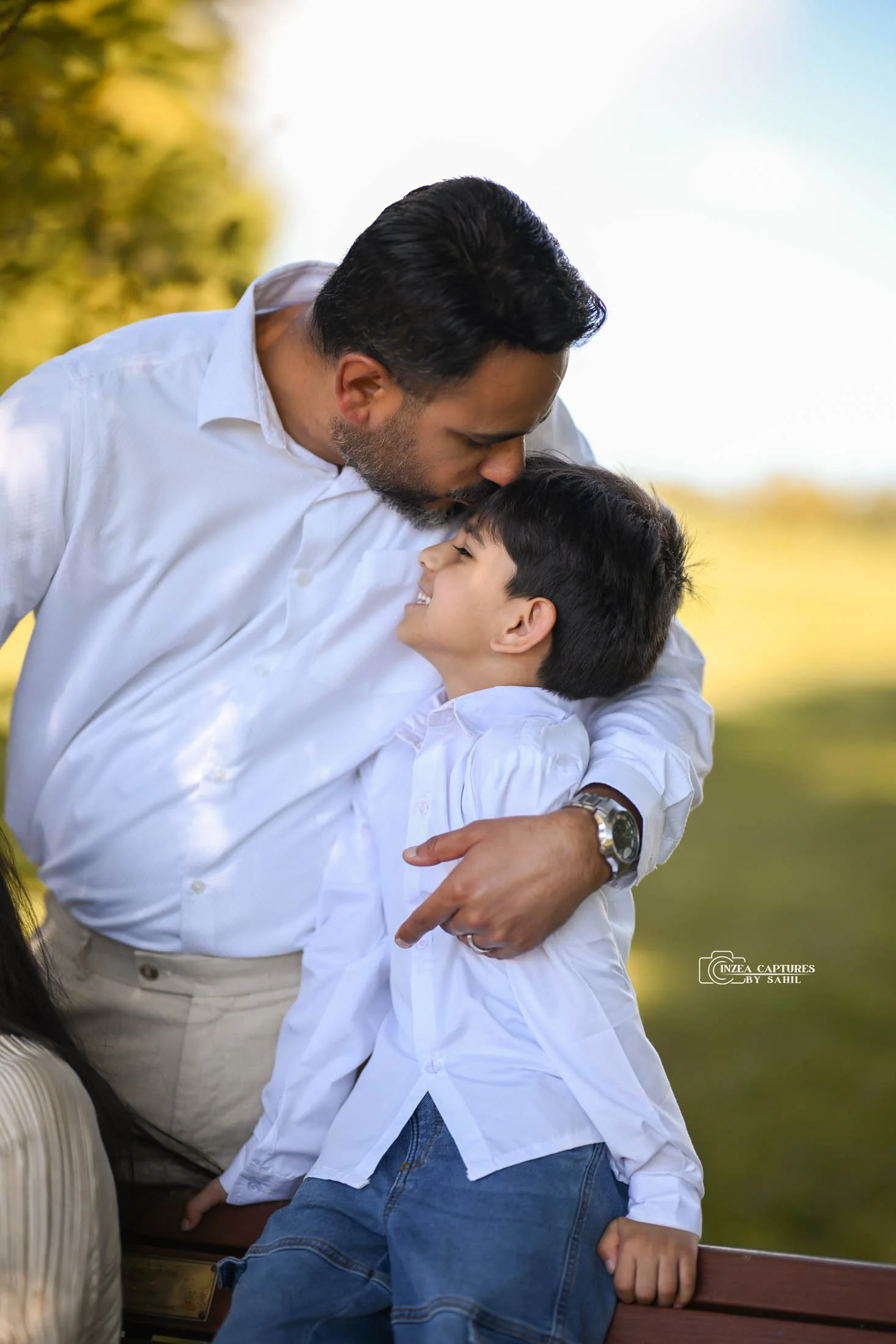 A man with dark hair and beard wearing a white shirt embraces a smiling young boy with dark hair also dressed in white in an outdoor setting with blurred trees in the background.