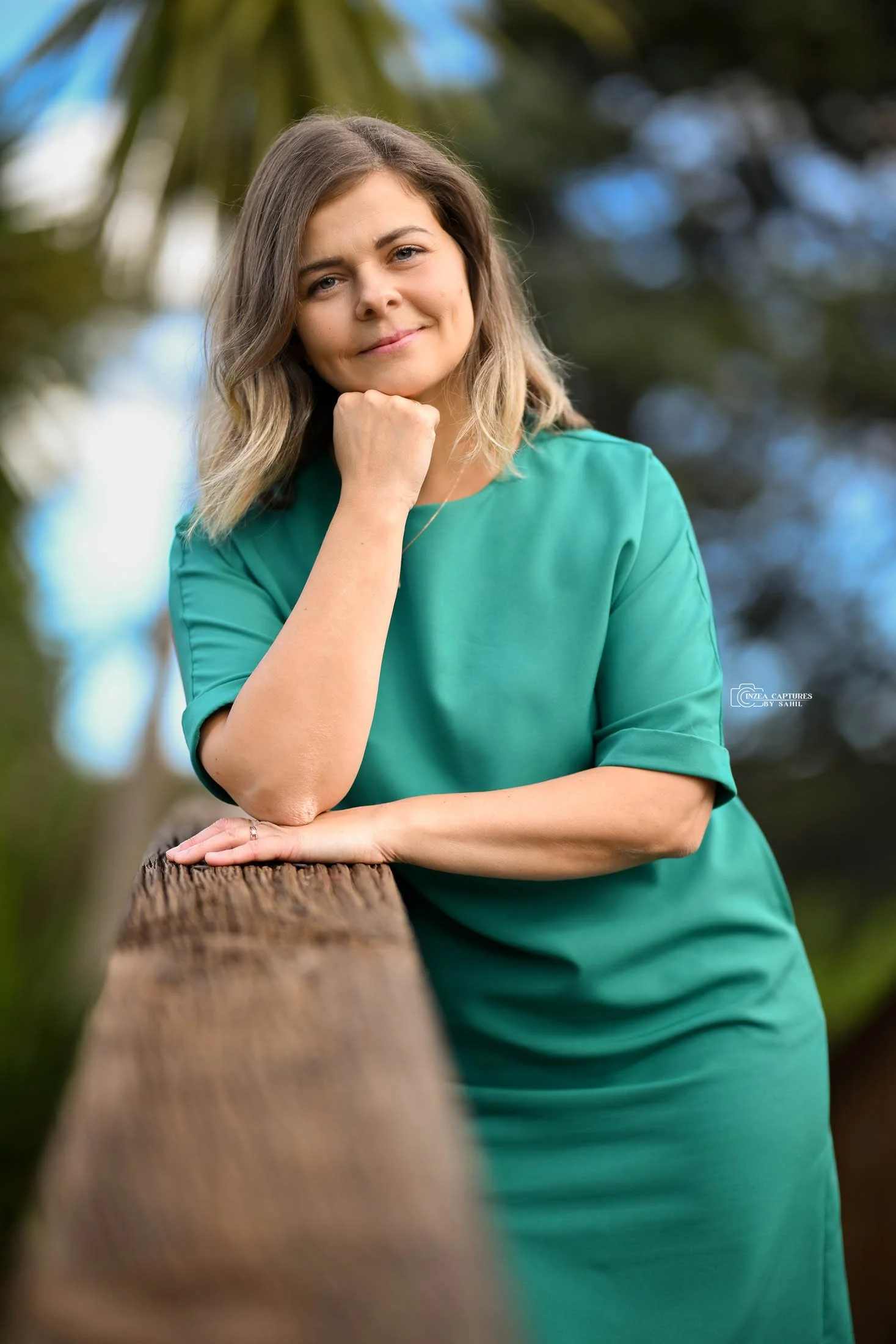 A woman in a teal dress leaning on a wooden railing outdoors, smiling gently at the camera, with blurred trees and sky in the background.