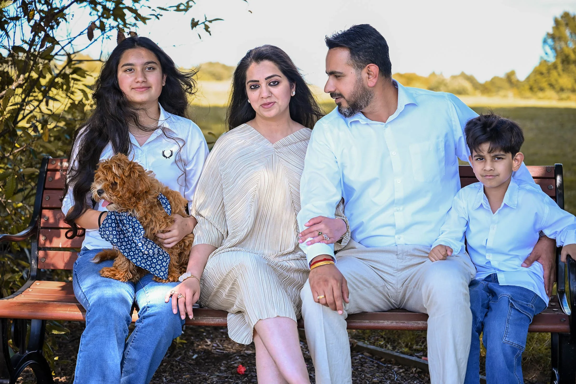 A family of five sitting on a park bench outdoors, with a dog in the girl's lap, surrounded by greenery and blue sky.