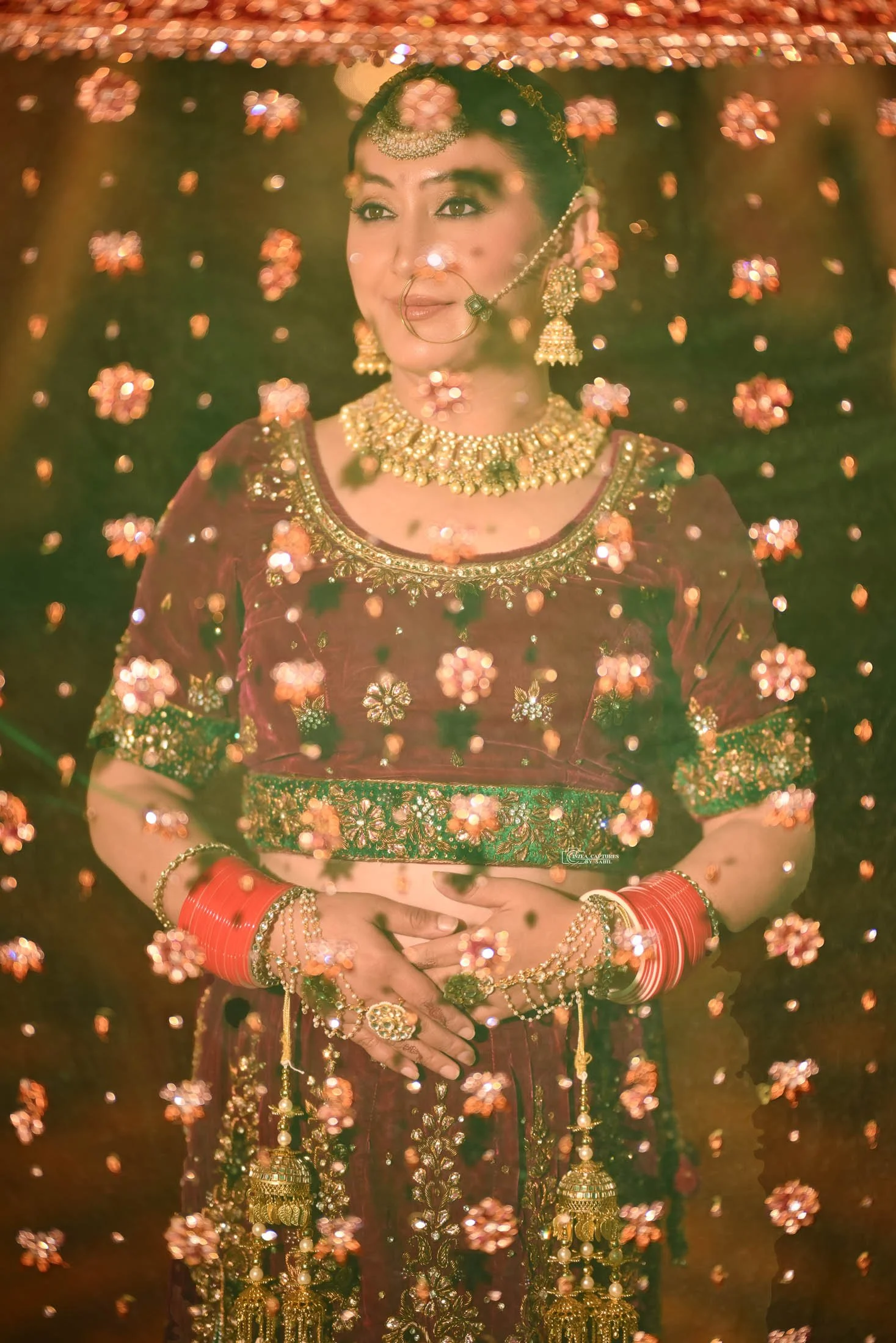 A woman dressed in traditional Indian bridal attire, adorned with gold jewelry, red bangles, and intricate embroidery, standing behind a decorative curtain of pink and gold flowers.