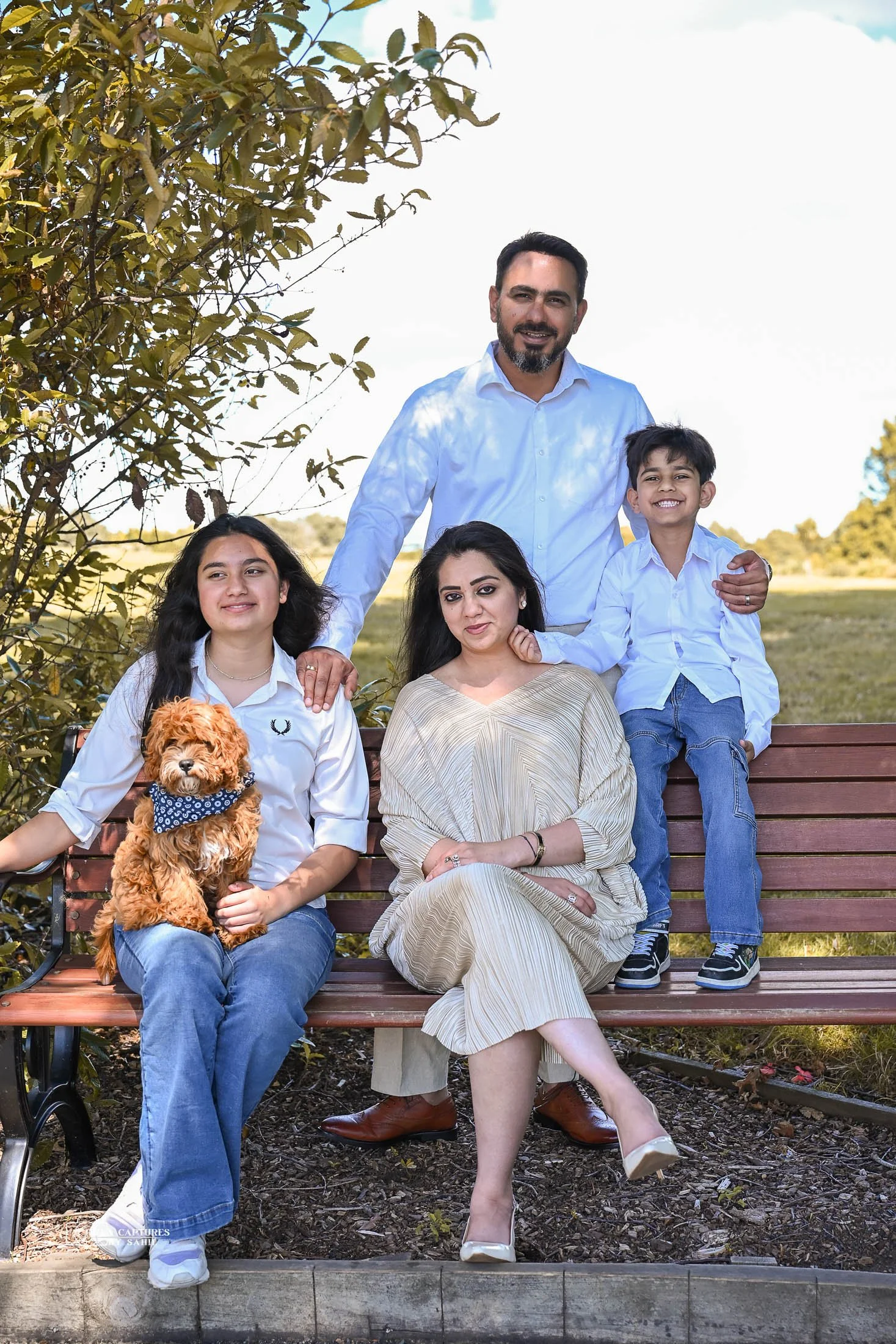 A family of five with a small dog sitting on a park bench outdoors during daytime, surrounded by greenery and a blue sky.