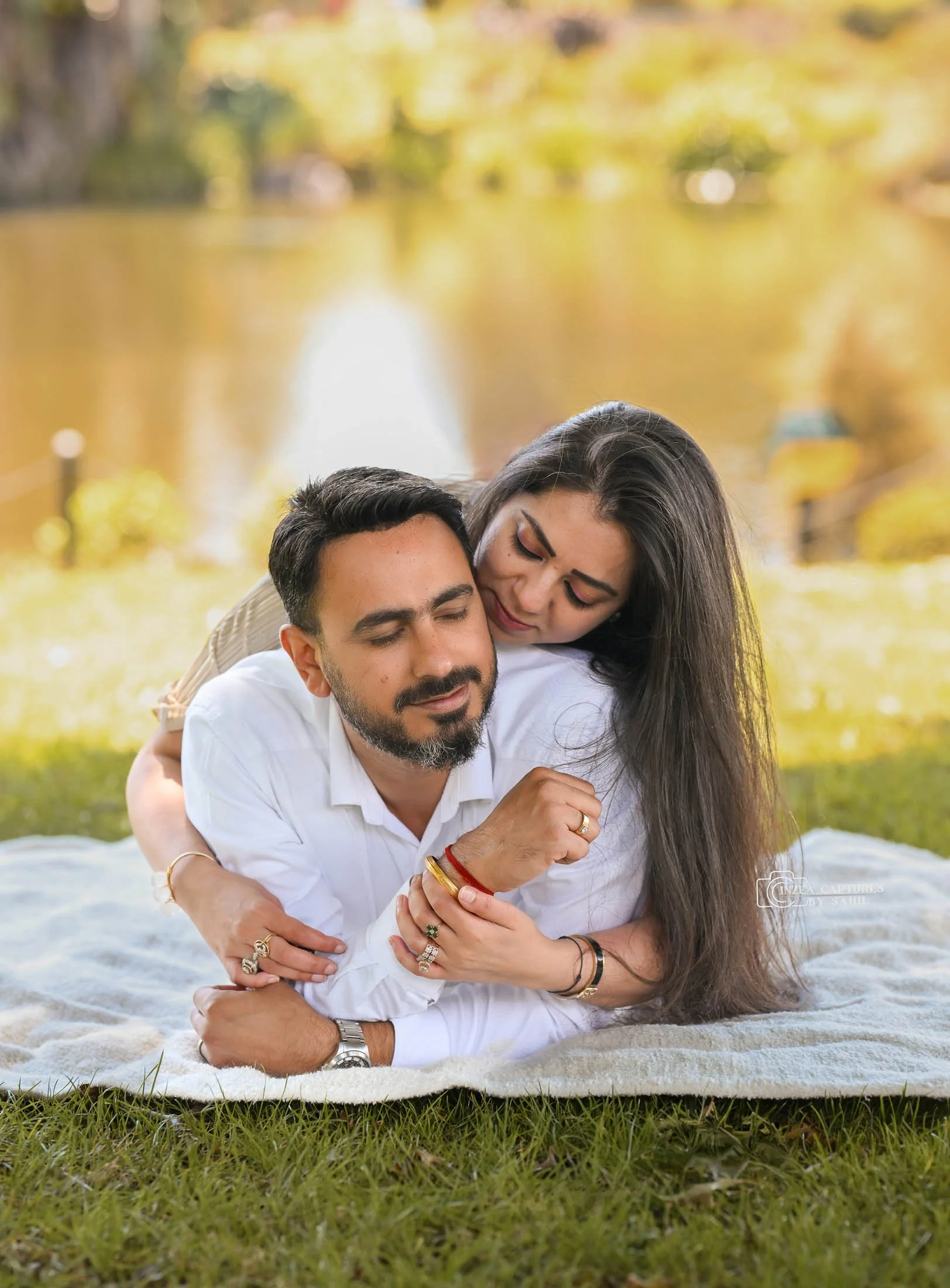 A couple lying on a blanket near a pond, enjoying a peaceful moment together in a park during autumn.
