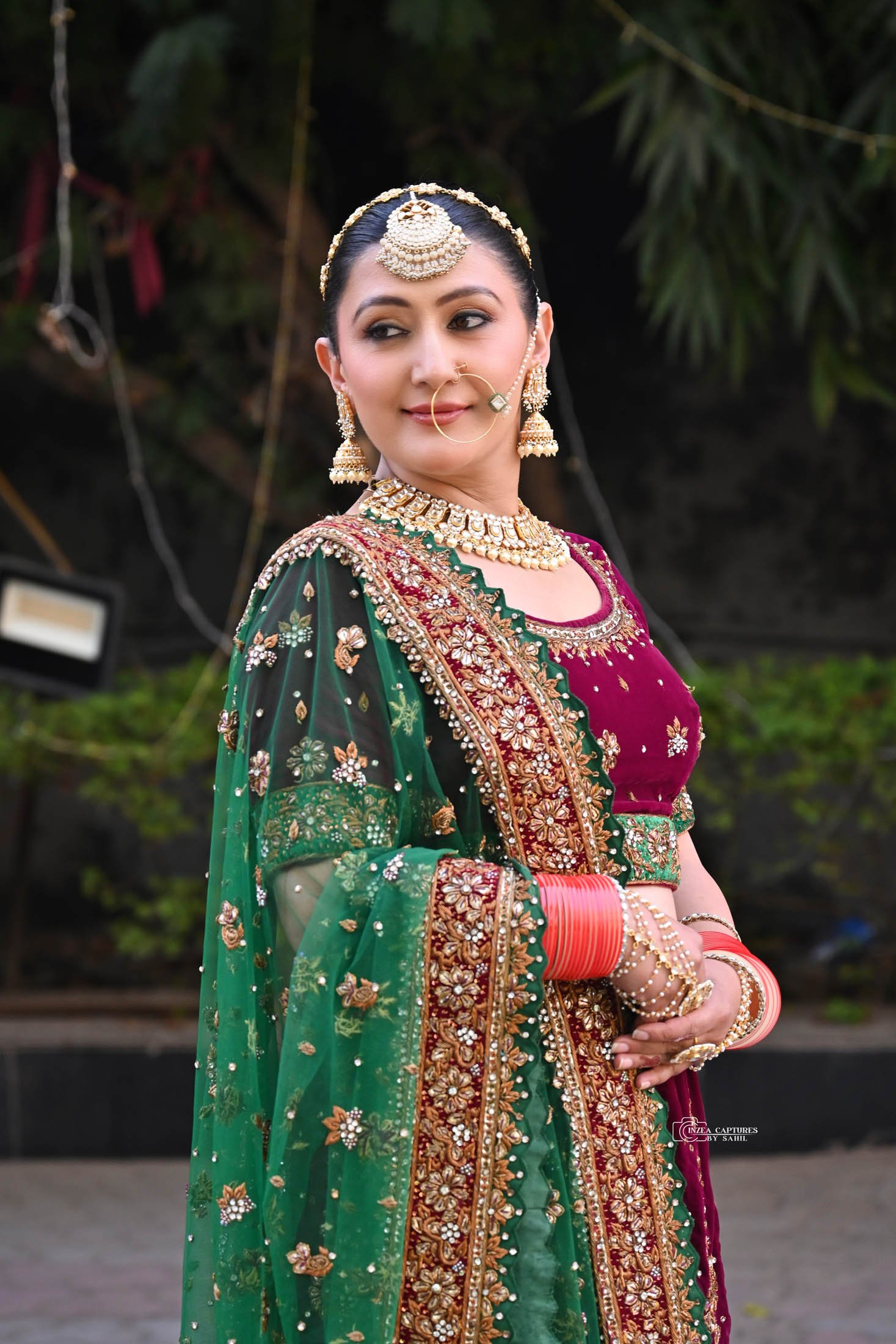 A woman in traditional Indian bridal attire, featuring a maroon velvet blouse, green and gold heavily embroidered dupatta, and traditional jewelry including a maang tikka, large earrings, nose ring, choker, and bangles, standing outdoors with trees i