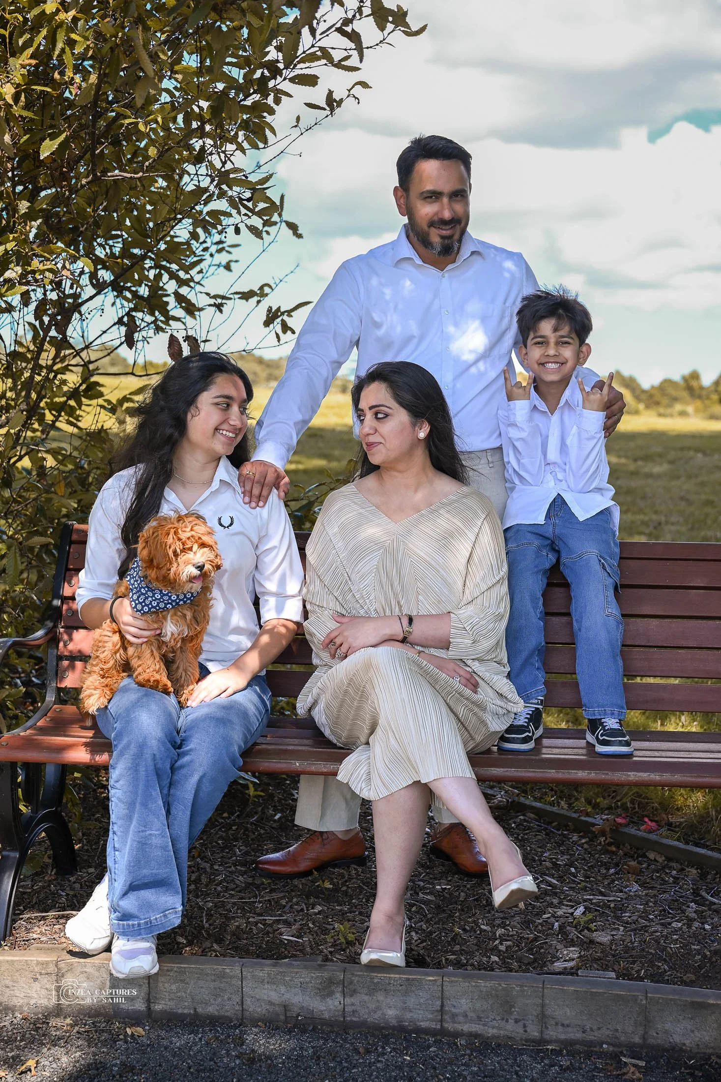 A family of five, including a woman, a man, two children, and a dog, sitting and standing on a park bench outdoors during daytime with a cloudy sky and greenery in the background.