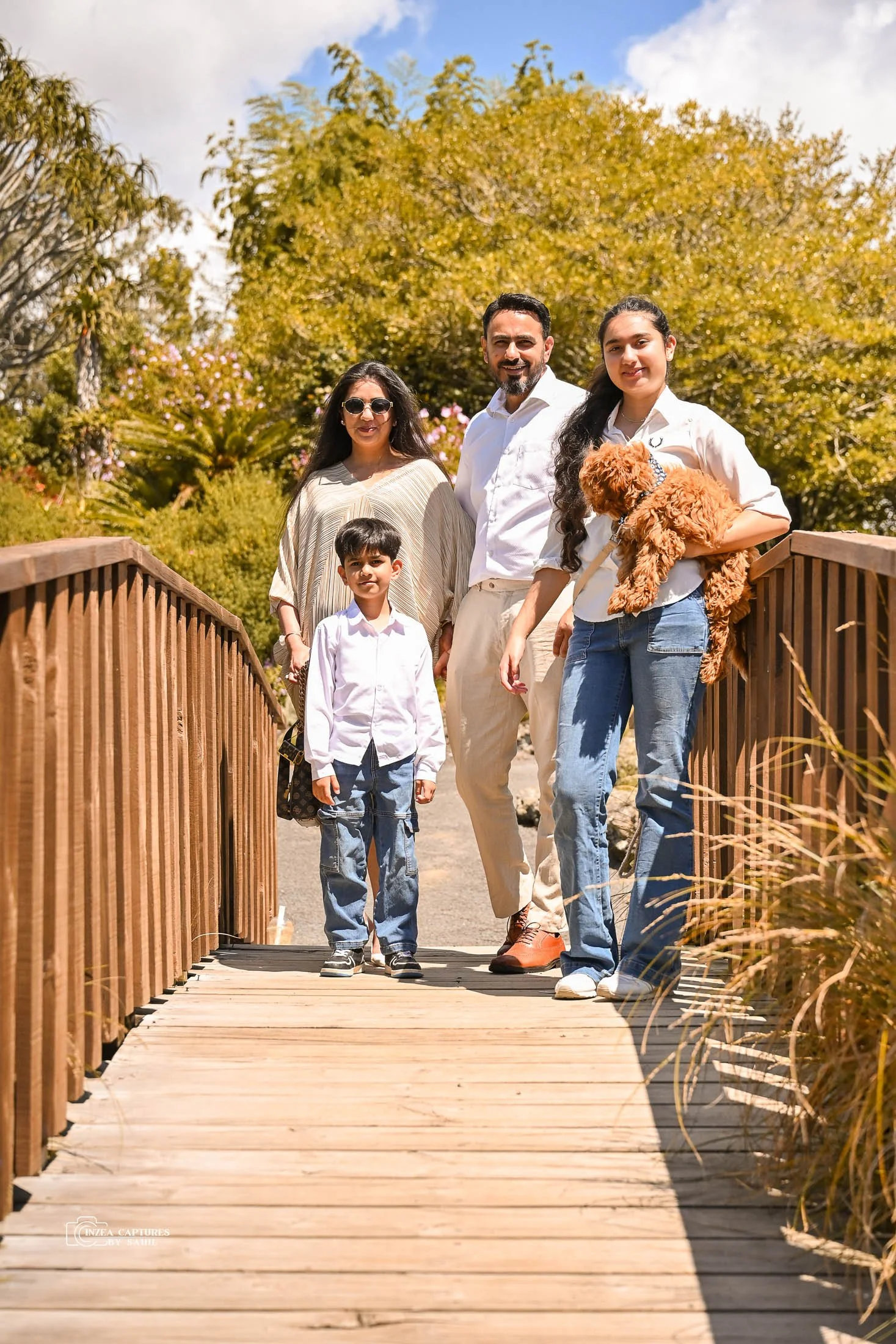 A family of five, including two children and a dog, standing on a wooden bridge outdoors with trees and greenery in the background on a sunny day.