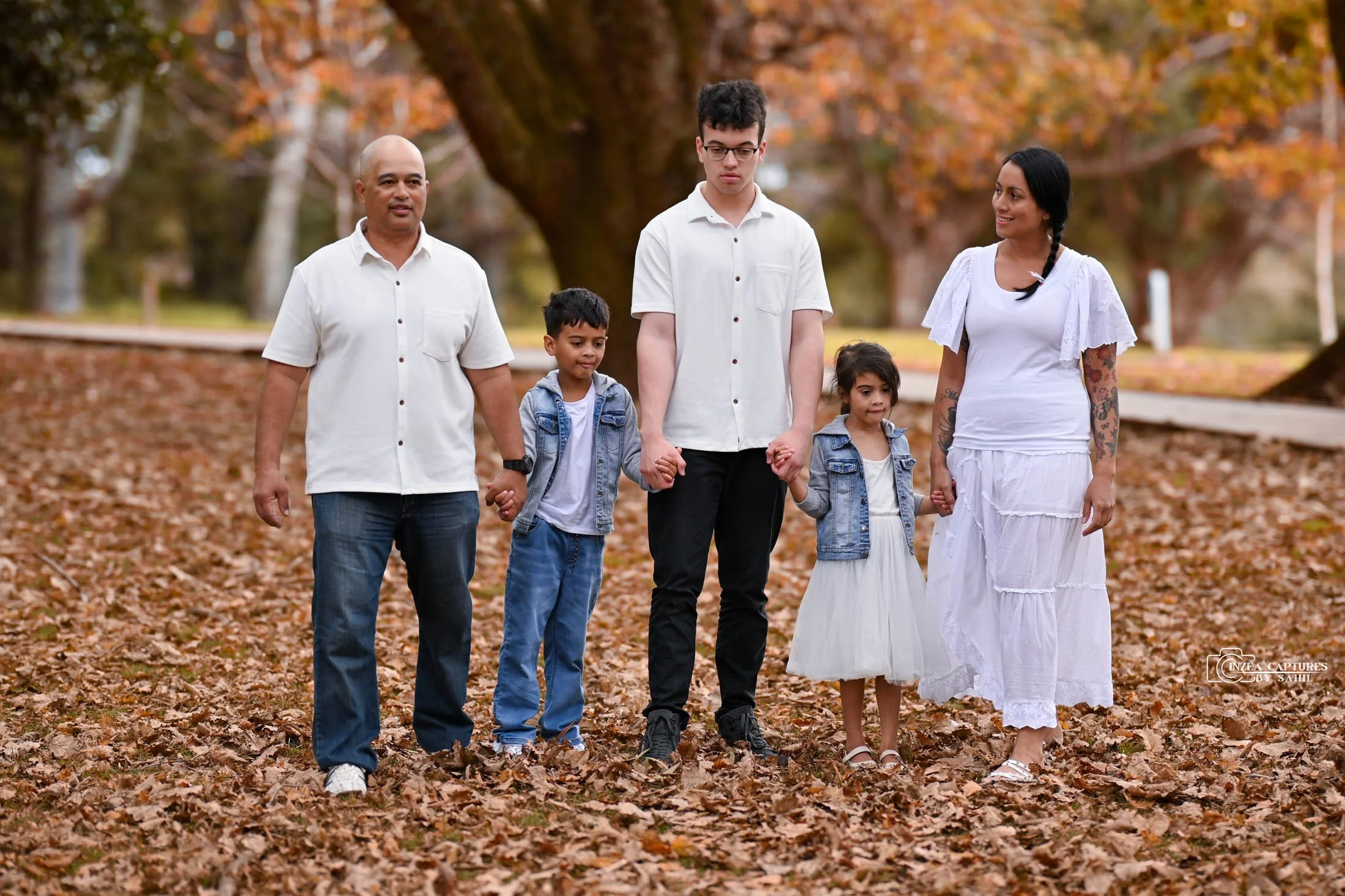 A multigenerational family of six walking hand-in-hand through a park covered with fallen autumn leaves. The background features large trees with orange and brown foliage.