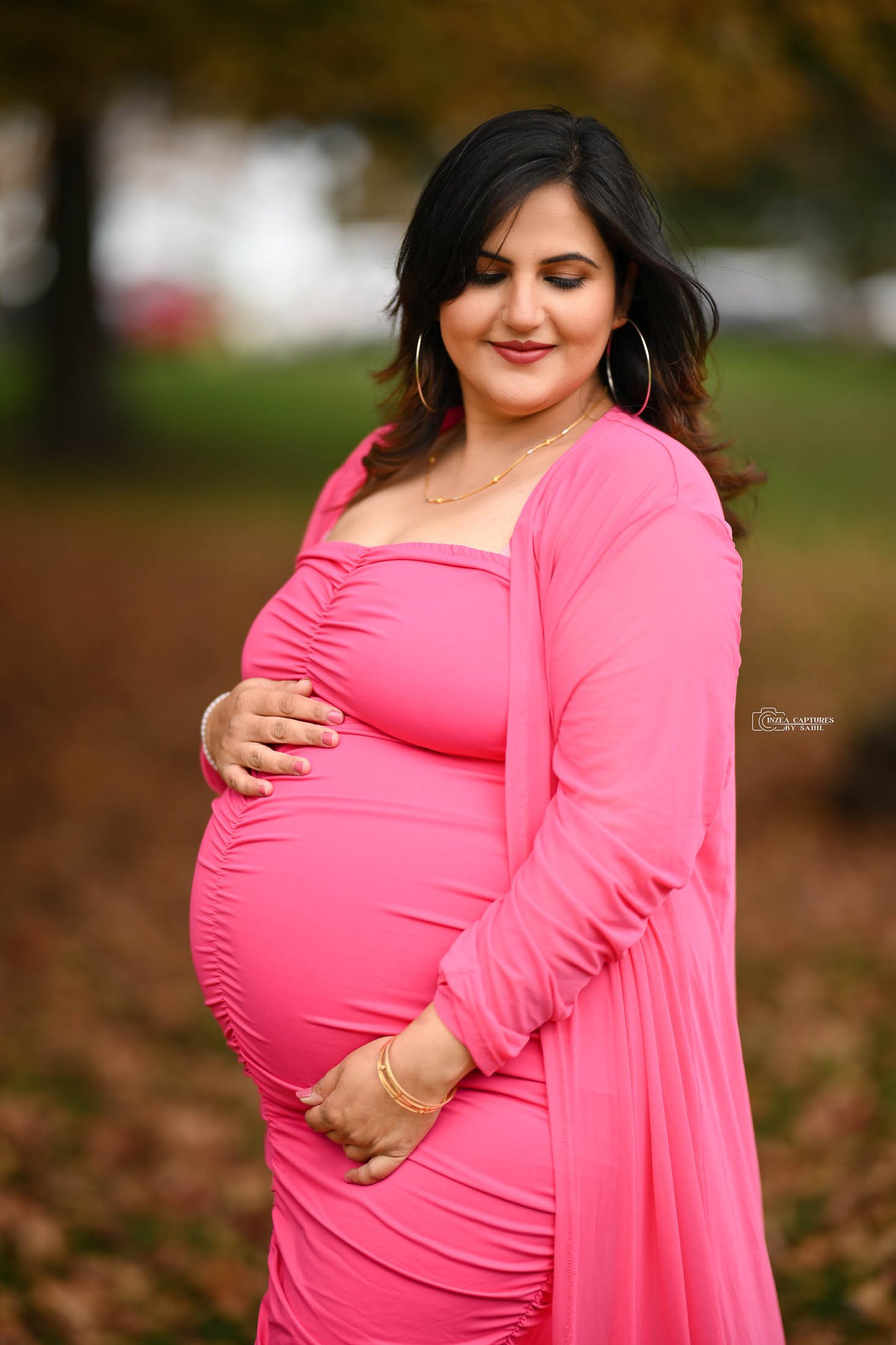 A pregnant woman in a pink dress and matching scarf stands outdoors, with trees and autumn foliage in the background, holding her belly and smiling gently.