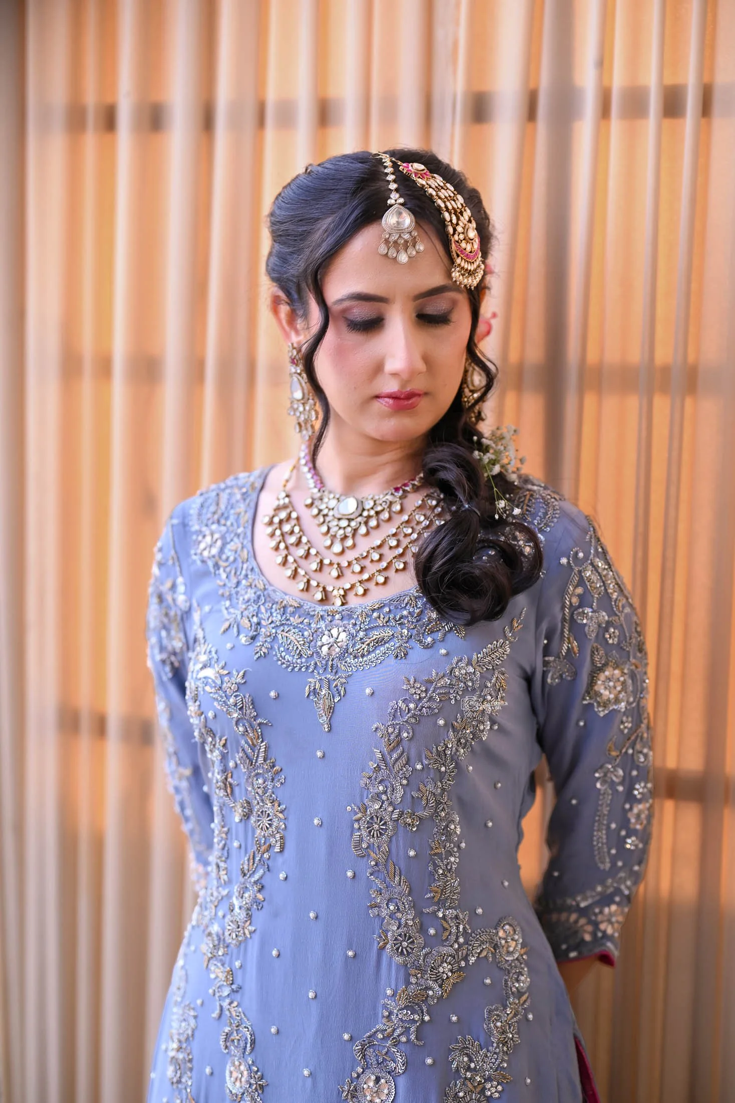 Woman in traditional Indian attire with elaborate jewelry and embroidery, standing in front of a curtain with her eyes closed.