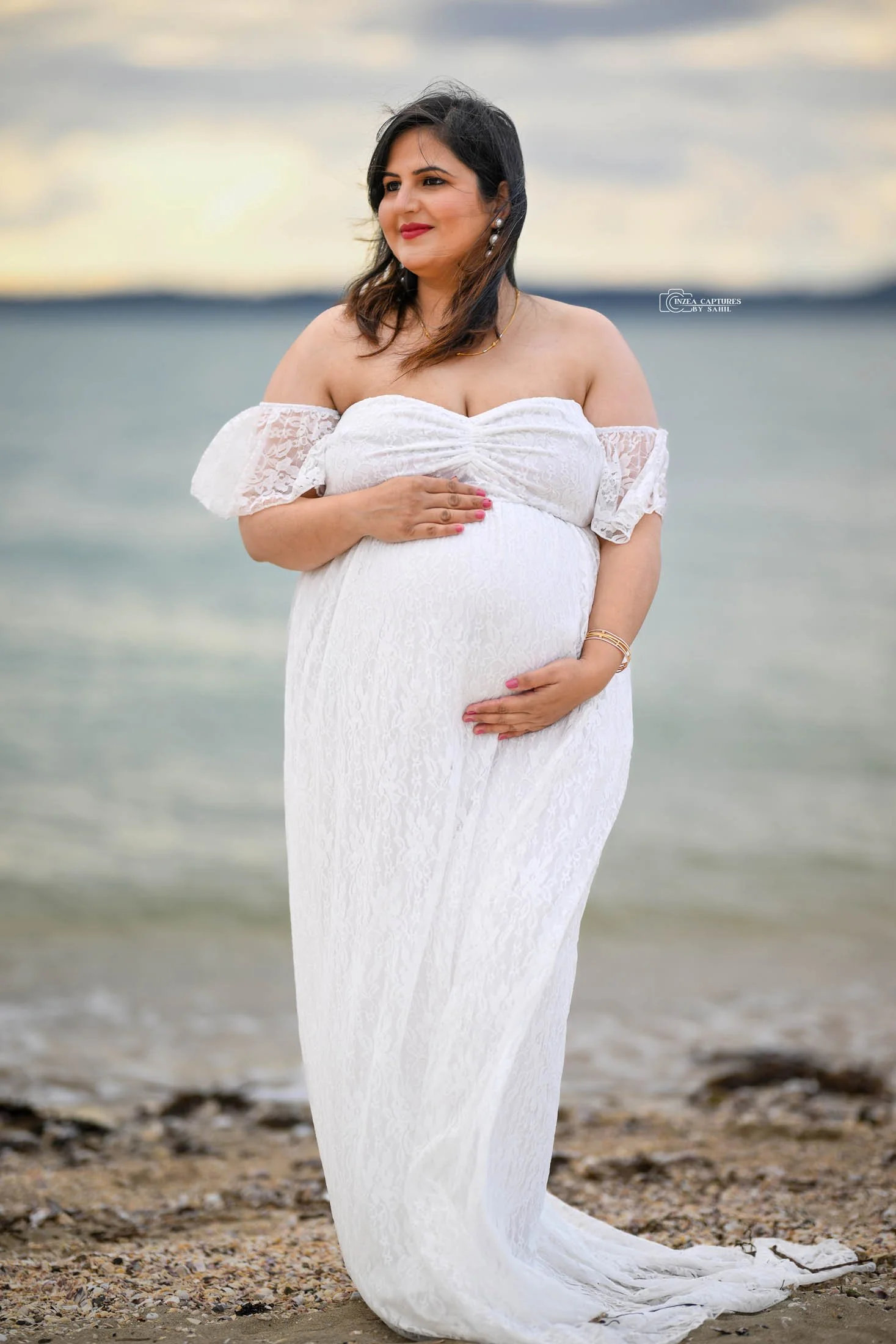 Pregnant woman in a white lace off-shoulder dress standing on a beach, holding her baby bump, with a blurred ocean and cloudy sky in the background.