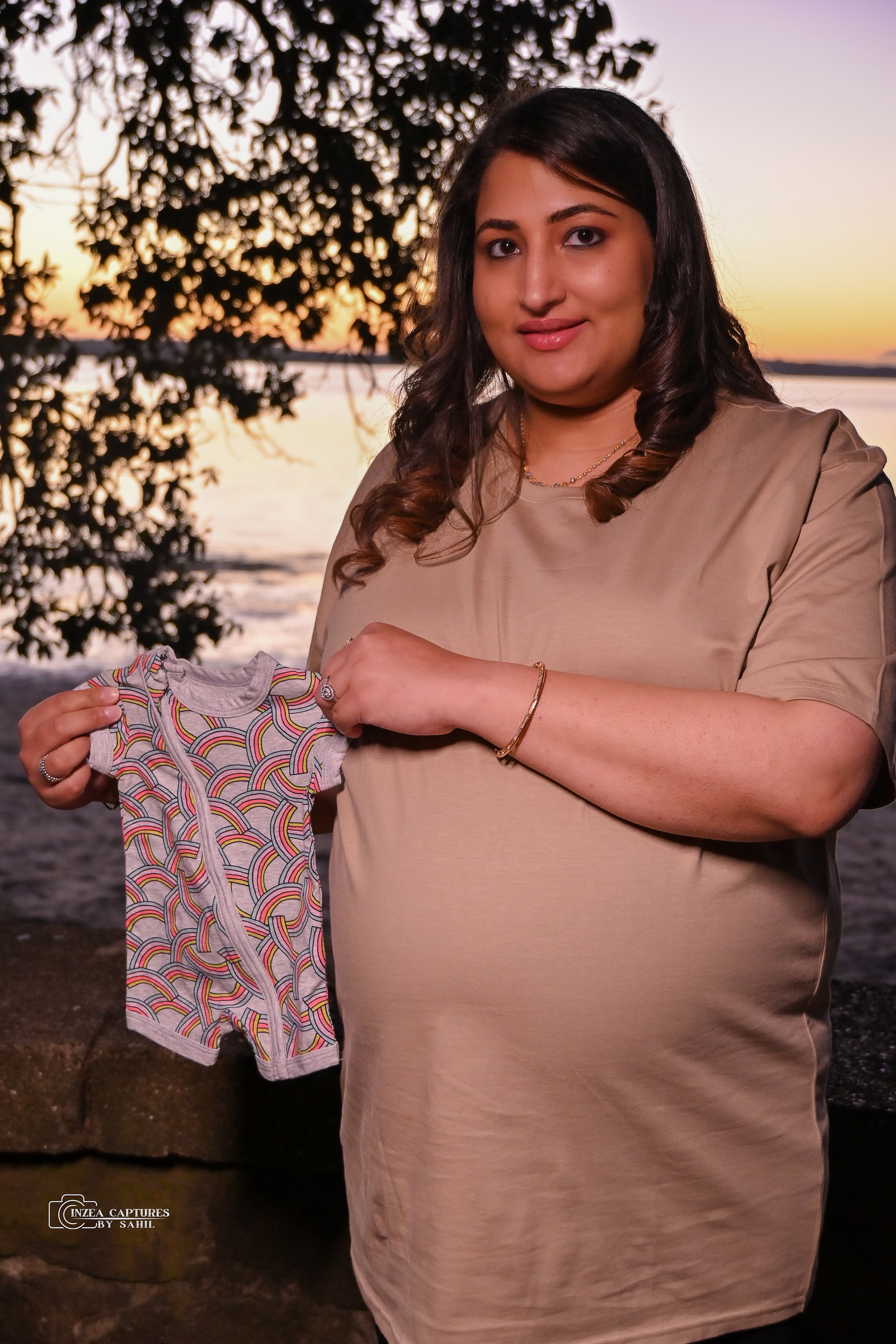 A woman standing outdoors near water during sunset, holding a colorful baby onesie with rainbow patterns.