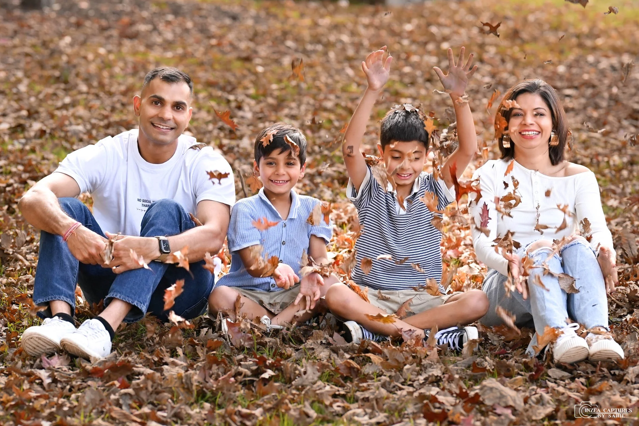 Family of four sitting on a bed of fallen autumn leaves outdoors, playing with leaves and smiling. The group includes a father, mother, and two young boys, enjoying a fall day.