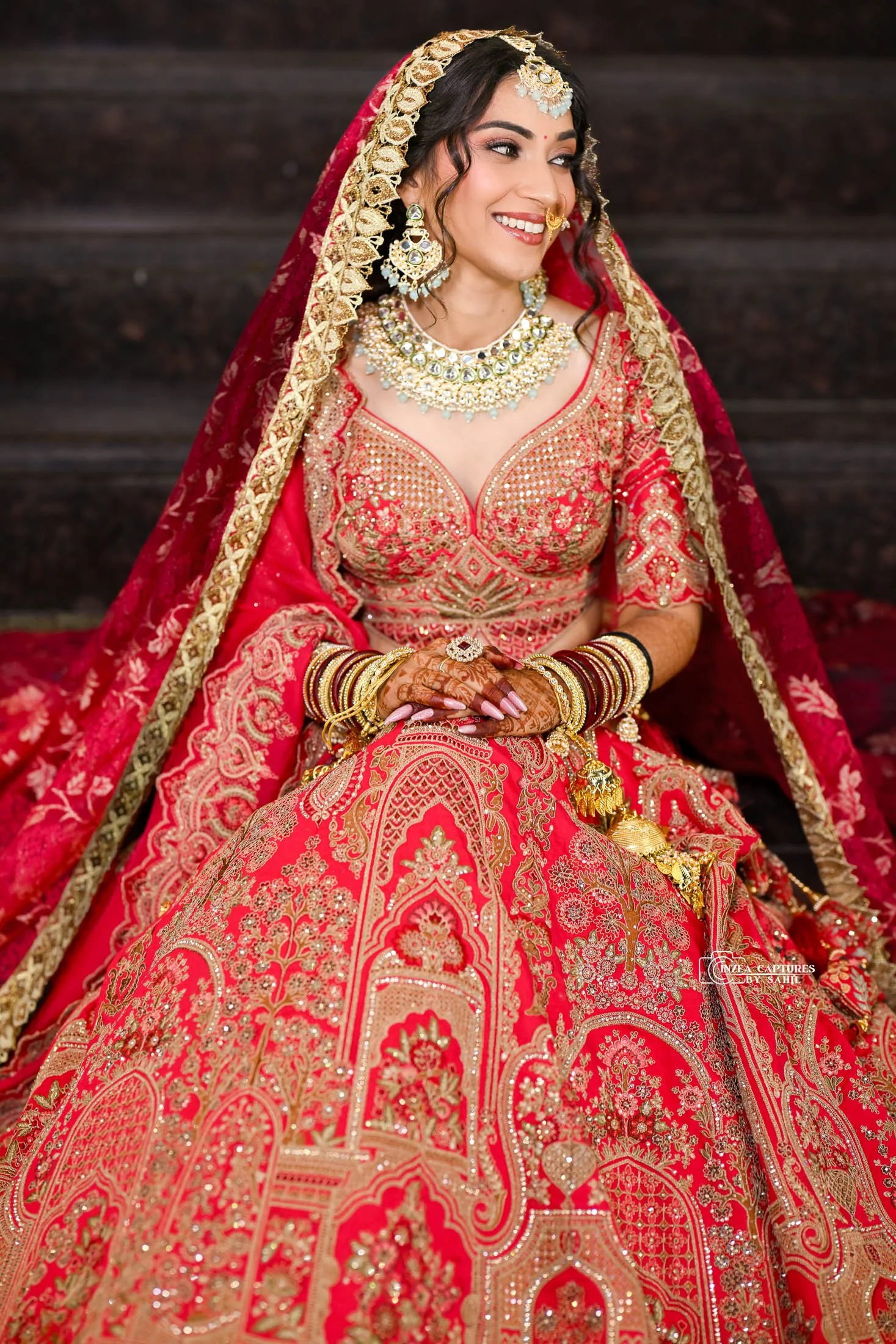A woman dressed in traditional Indian bridal attire, wearing a red and gold embroidered saree, ornate jewelry including a necklace, earrings, and headpiece, and adorned with bangles and henna, sitting on steps and smiling.