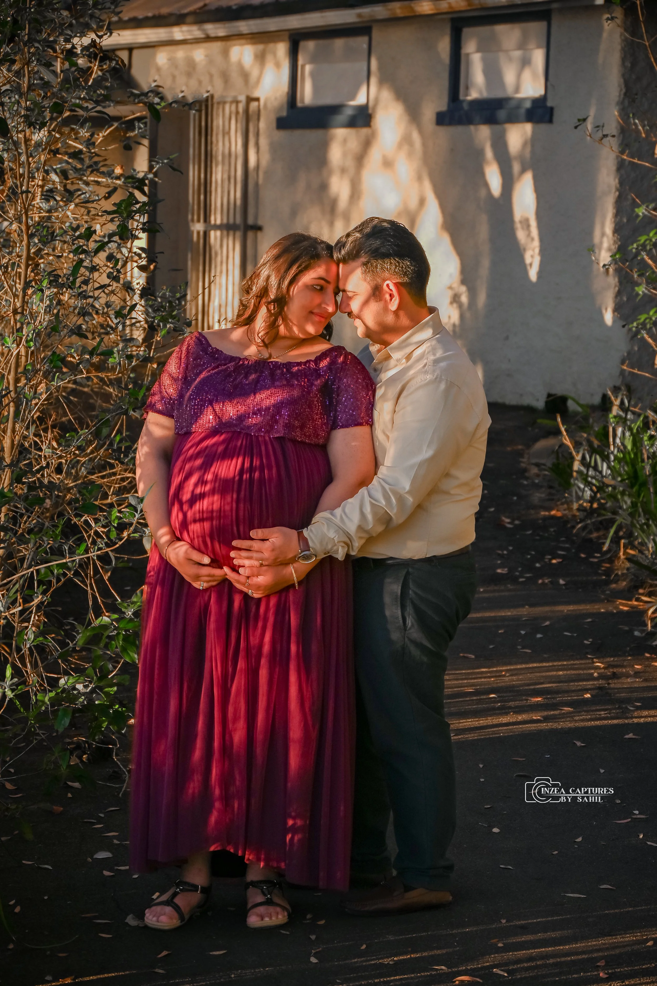 A pregnant woman and a man in outdoor setting, touching foreheads and holding her baby bump, during sunset with shadows and warm light.