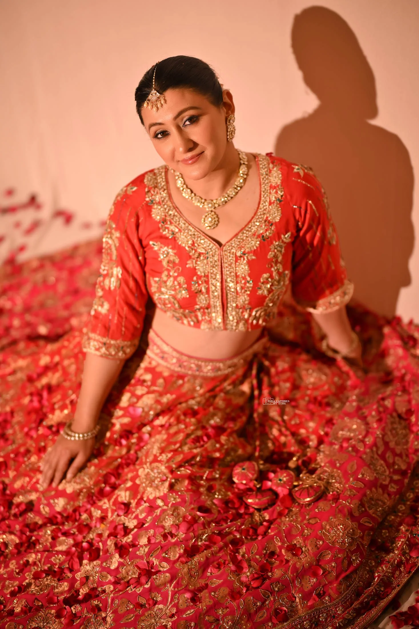 A woman in traditional Indian bridal attire sitting on a decorated surface, wearing a red embroidered lehenga with gold accents, jewelry, and a bindi.
